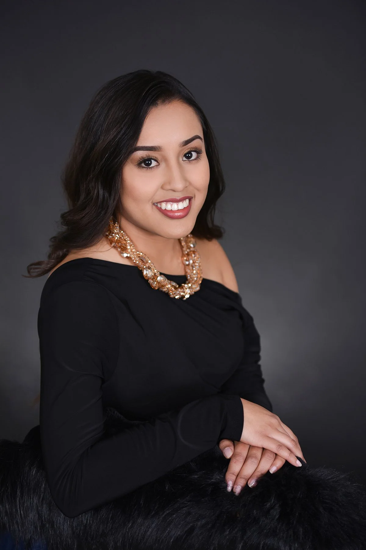 A woman with black hair and a warm smile, wearing a black off-shoulder top and a gold beaded necklace, sitting with her hands clasped, against a dark gray background.