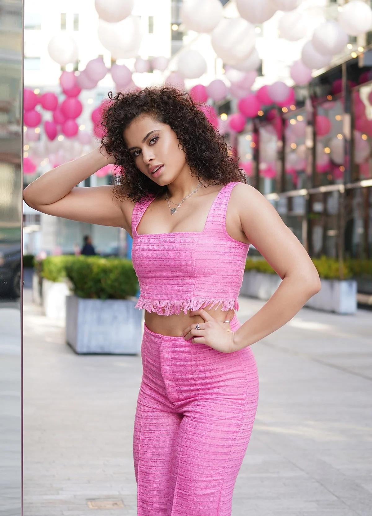 A young woman with curly hair wearing a pink sleeveless crop top and matching pink pants, standing outdoors with pink and white balloon decorations hanging above her.
