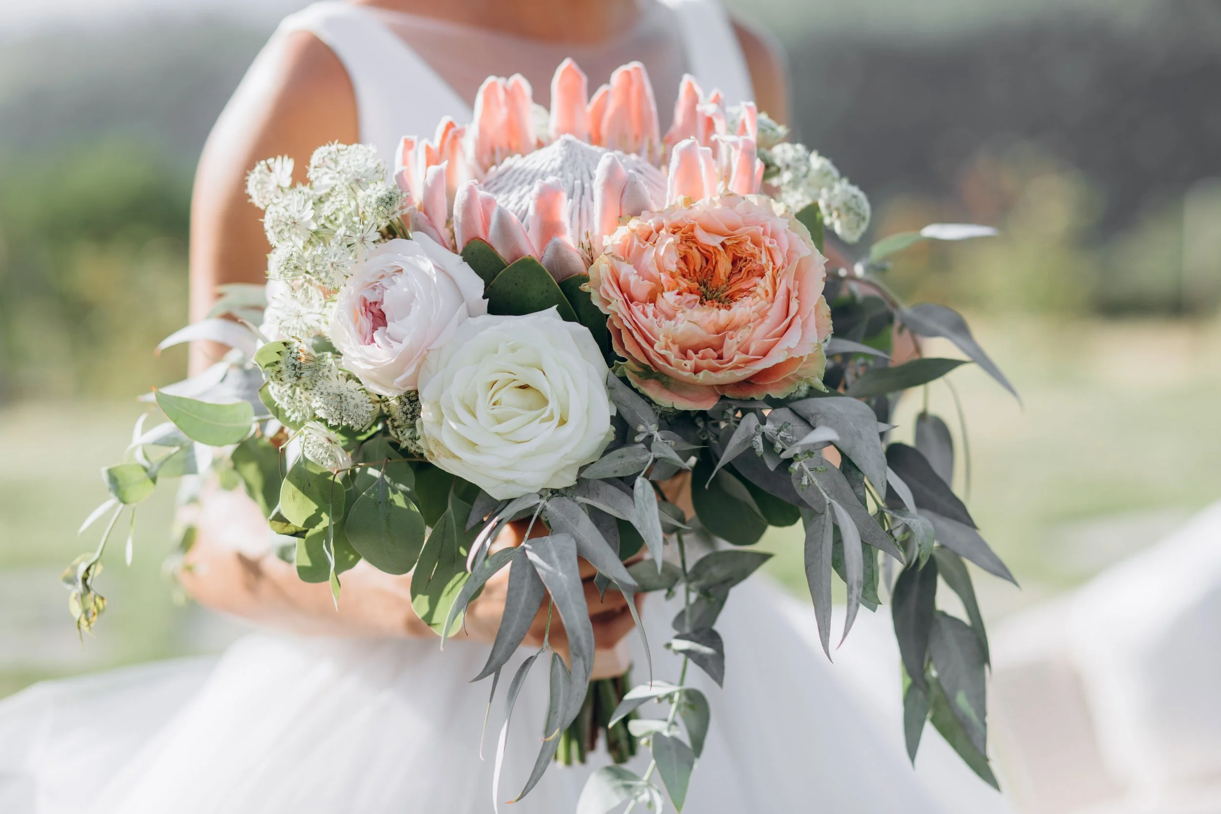 A person holding a bouquet of flowers including roses, peonies, and other blossoms in soft pink and white colors with green and gray foliage, outdoors with blurred greenery in the background.