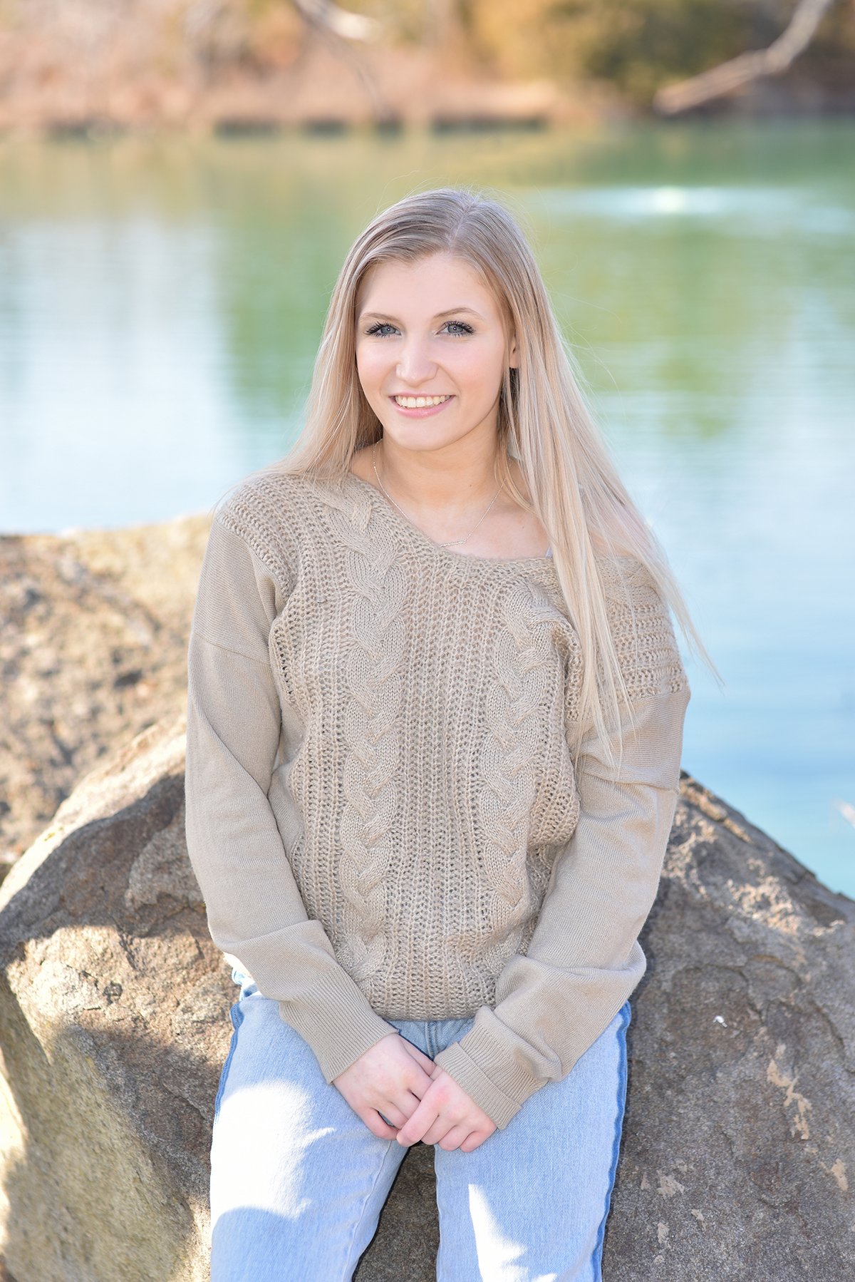 A young woman with long blonde hair sitting on a rock near a body of water, wearing a beige knit sweater and light blue jeans, smiling at the camera.