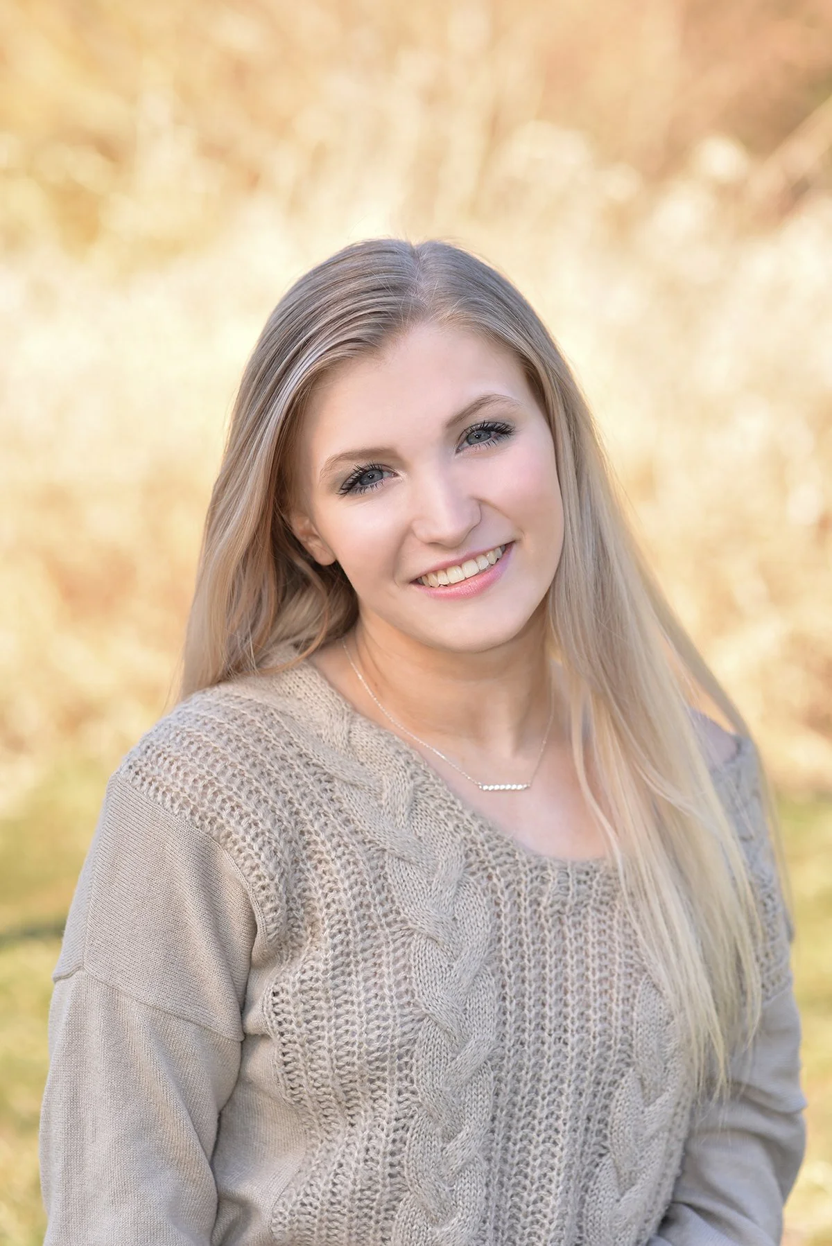 Portrait of a young woman with long blonde hair, smiling, wearing a beige sweater and a silver necklace, outdoors with a blurred yellow foliage background.