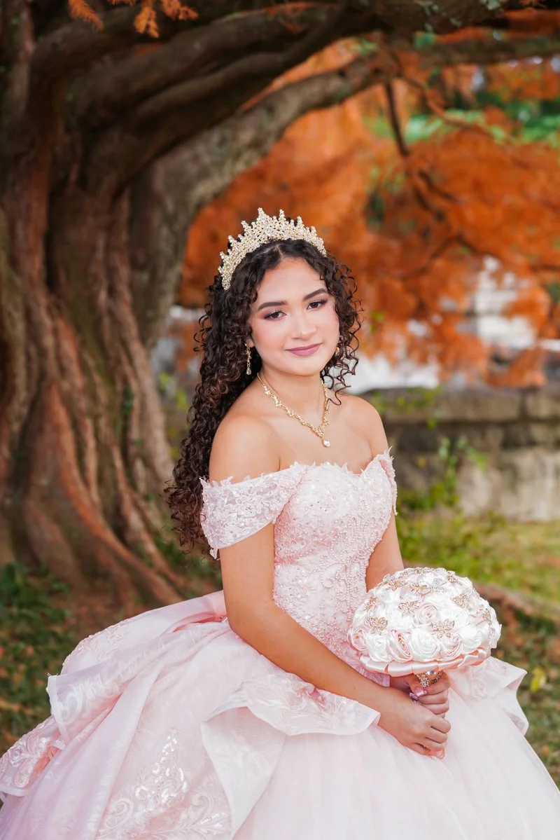 A young woman in a pink off-the-shoulder ball gown with lace and embroidery, wearing a tiara and jewelry, holding a bouquet of roses, standing outdoors in front of a large tree with orange autumn leaves.