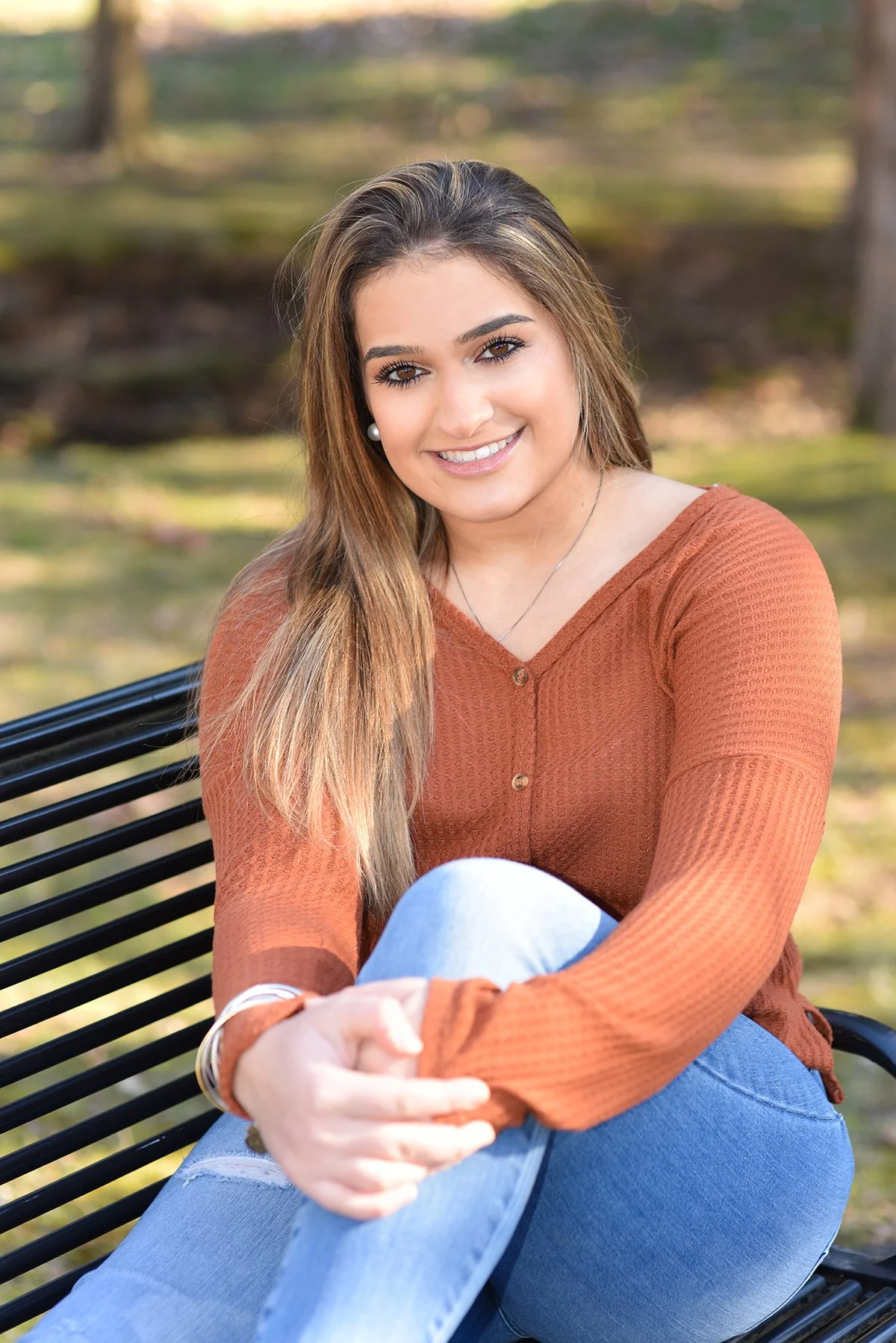 A smiling young woman with long hair and pearl earrings sitting on a park bench outdoors, wearing an orange long-sleeve shirt and light blue jeans.