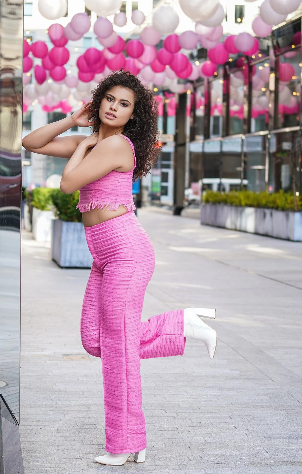 A woman with curly dark hair wearing a pink fringed top and matching wide-leg pants, white high-heeled boots, posing outdoors surrounded by pink and white paper lantern decorations.