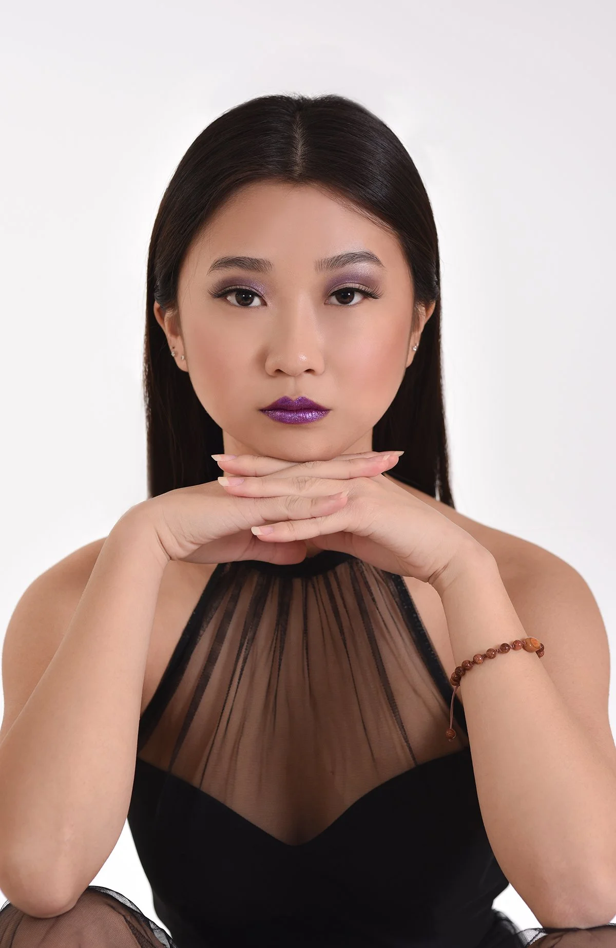 A woman with dark hair and purple lipstick, wearing a black sheer top, is posing with her hands under her chin against a plain white background.