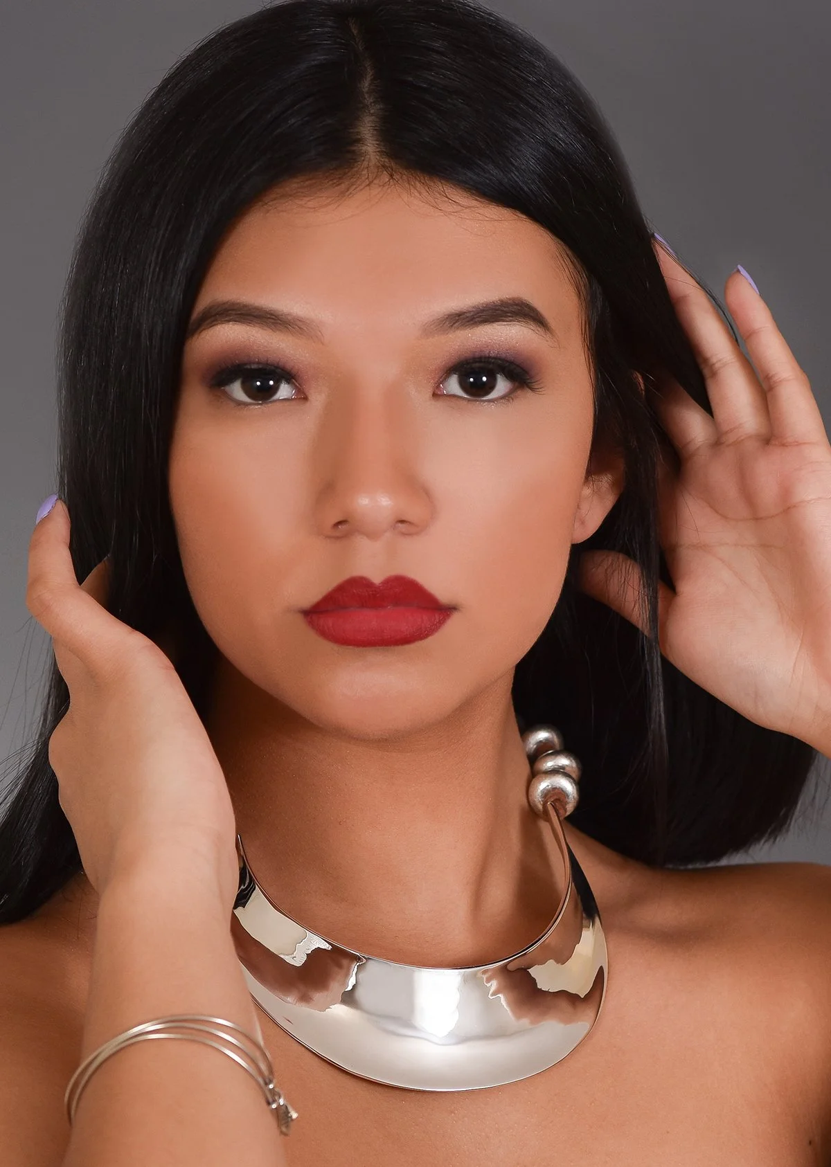 Close-up portrait of a young woman with dark hair, red lipstick, and silver jewelry, posing with hands near her face against a gray background.