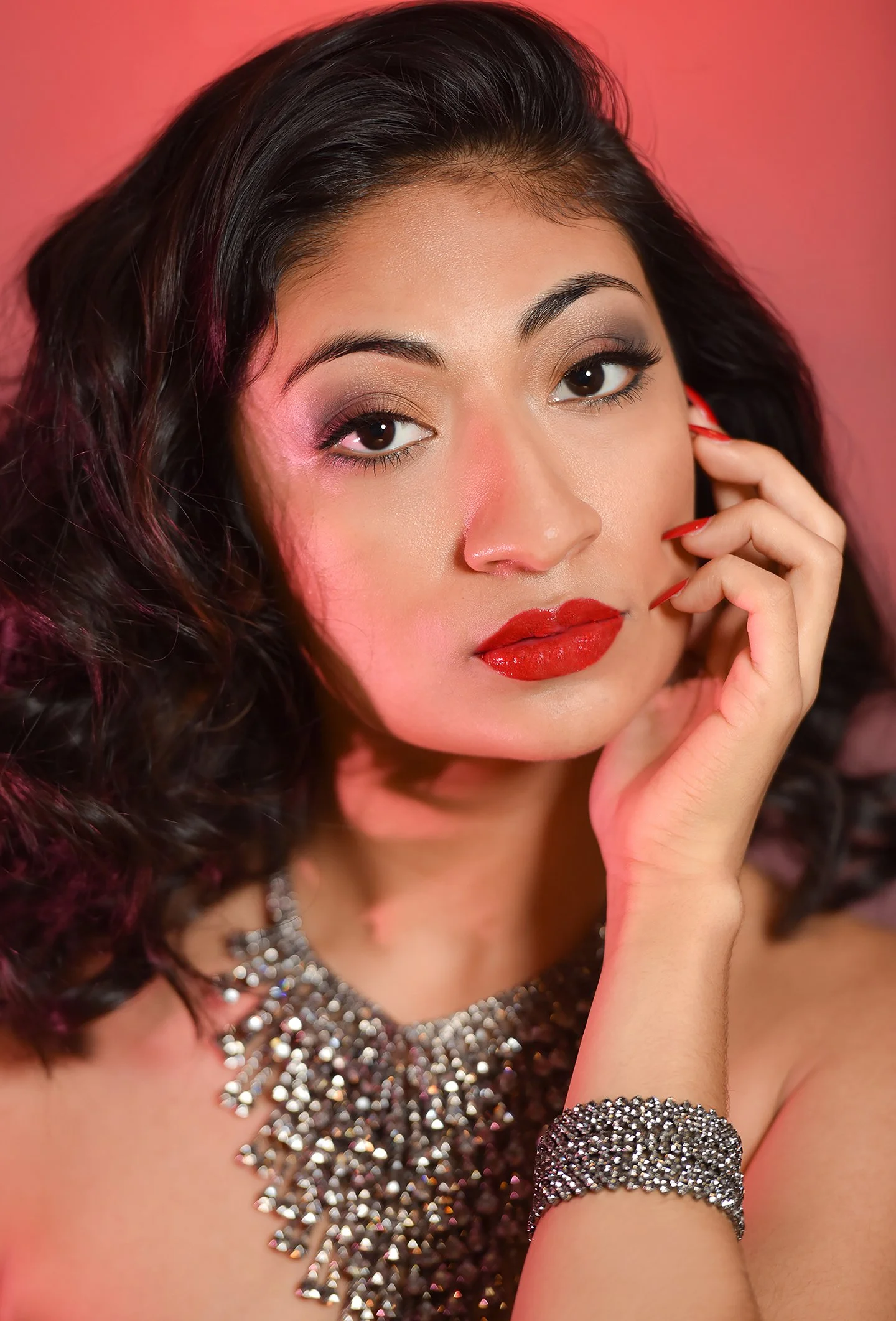 Close-up of a woman with dark curly hair, red lipstick, dark eye makeup, and a silver necklace and bracelet, against a pink background.