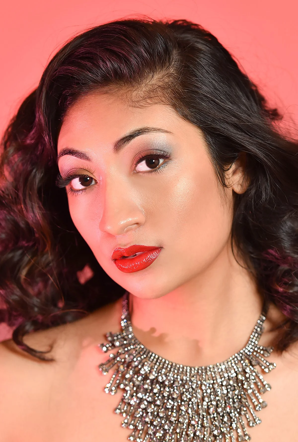 Close-up portrait of a woman with dark, curly hair, wearing bold makeup with silver eyeshadow and red lipstick, and a large, shiny, jeweled necklace against a pink background.