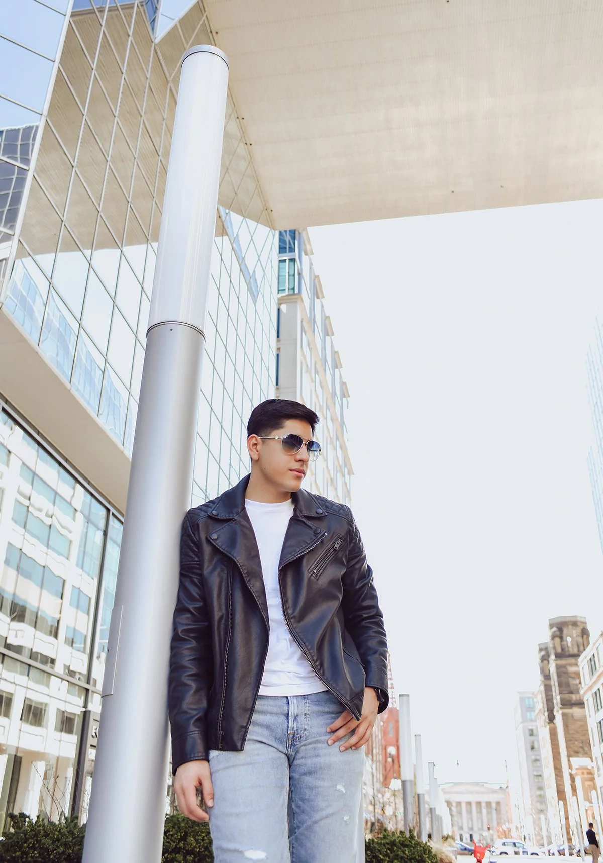 Young man with dark hair, wearing sunglasses, a black leather jacket, a white T-shirt, and light blue ripped jeans, standing outdoors in an urban area with modern glass buildings and a city skyline in the background.