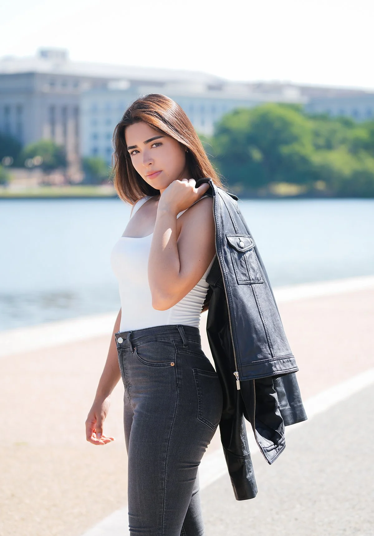 Young woman with shoulder-length brown hair, wearing a white tank top and black jeans, holding a black leather jacket over her shoulder, standing near a body of water with a cityscape in the background.
