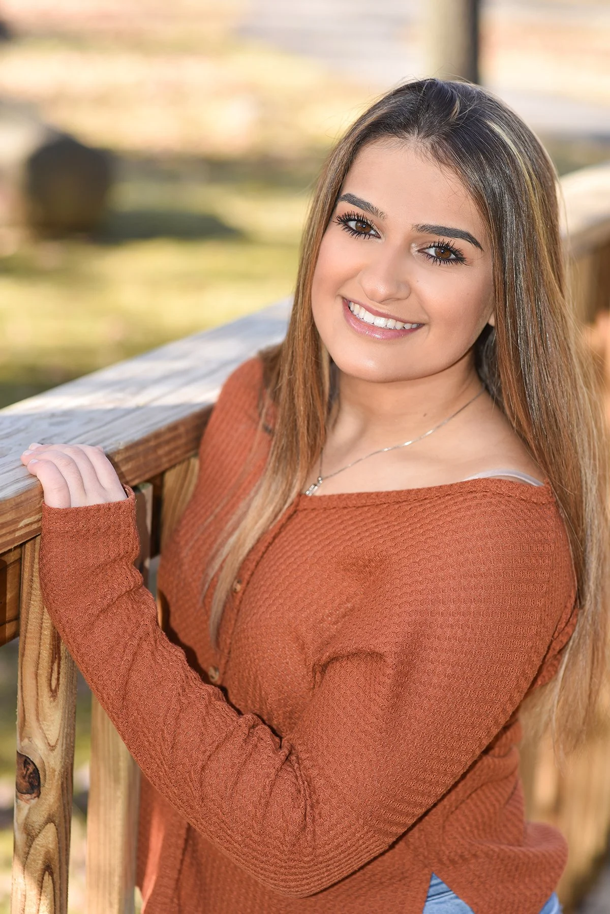 A young woman with long brown hair and brown eyes, smiling and looking at the camera, standing outdoors on a wooden fence with a blurred background of nature and sunlight.