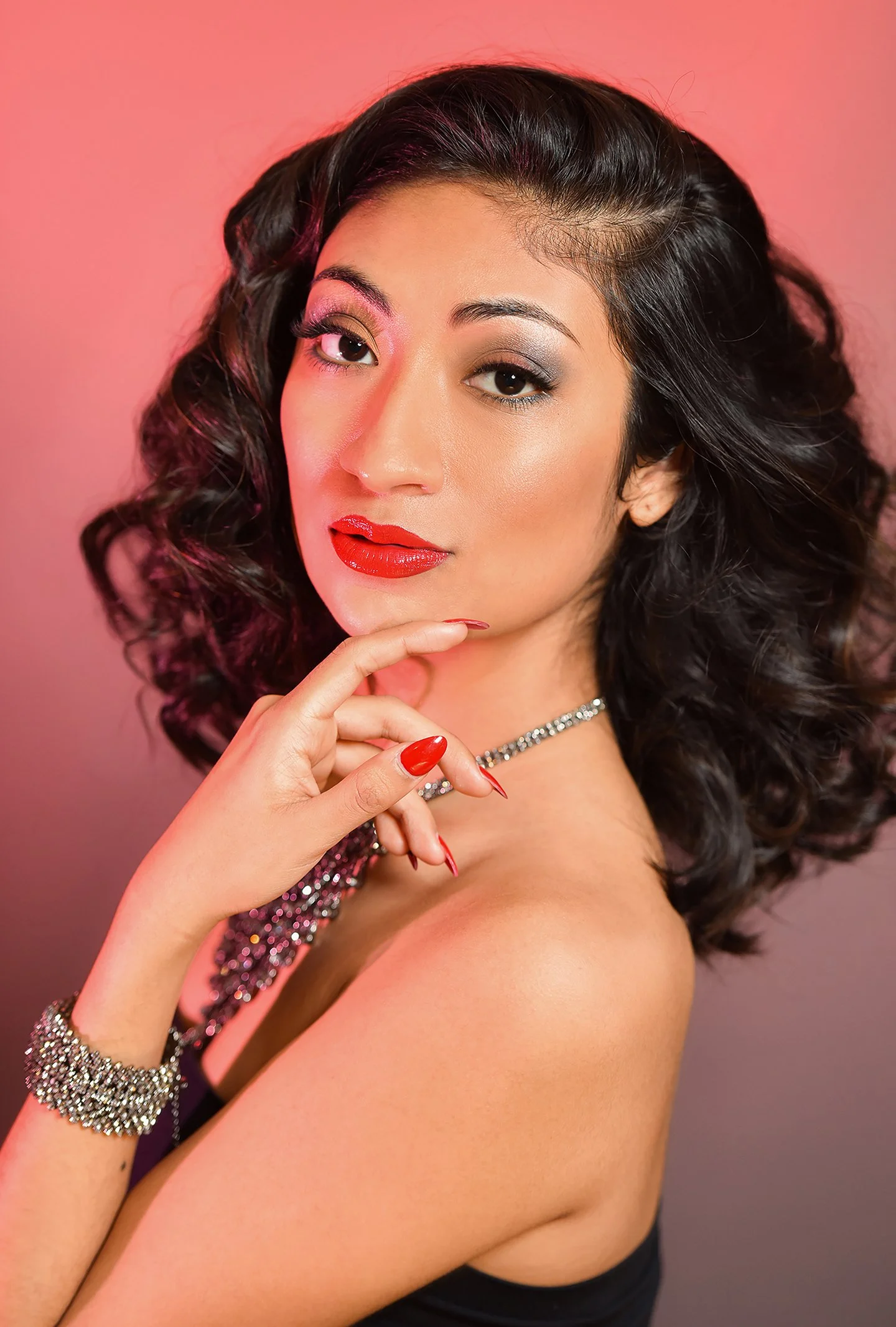 A woman with dark, curly hair styled in voluminous waves, wearing eye makeup with pink and silver eyeshadow, red lipstick, and a diamond necklace. She is touching her chin with her left hand, which has red nail polish, and is wearing a sparkly bracel