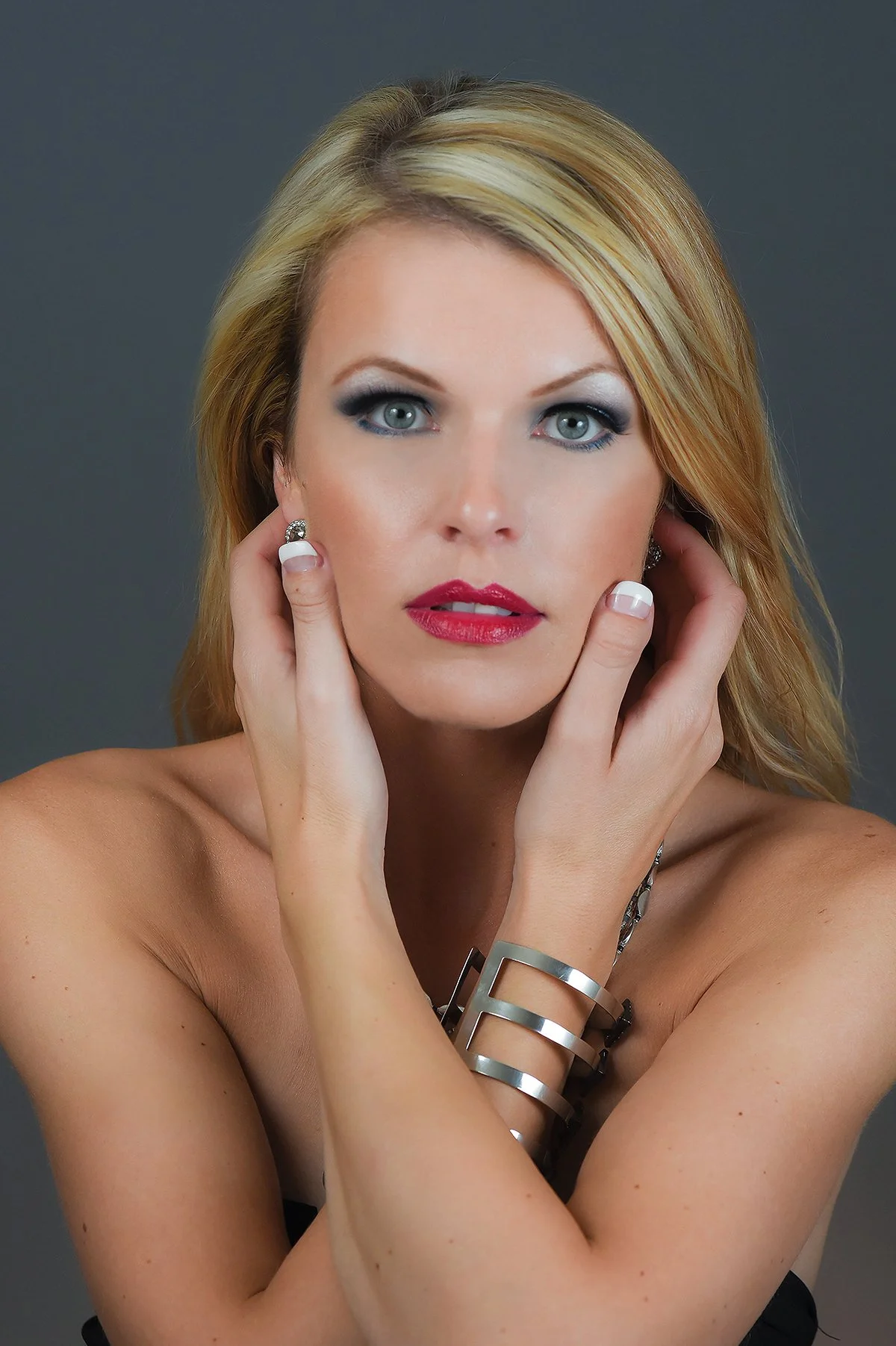 Portrait of a woman with blonde hair, blue eyes, and makeup, touching her face with her hands, wearing silver jewelry and a black strap top, with a gray background.