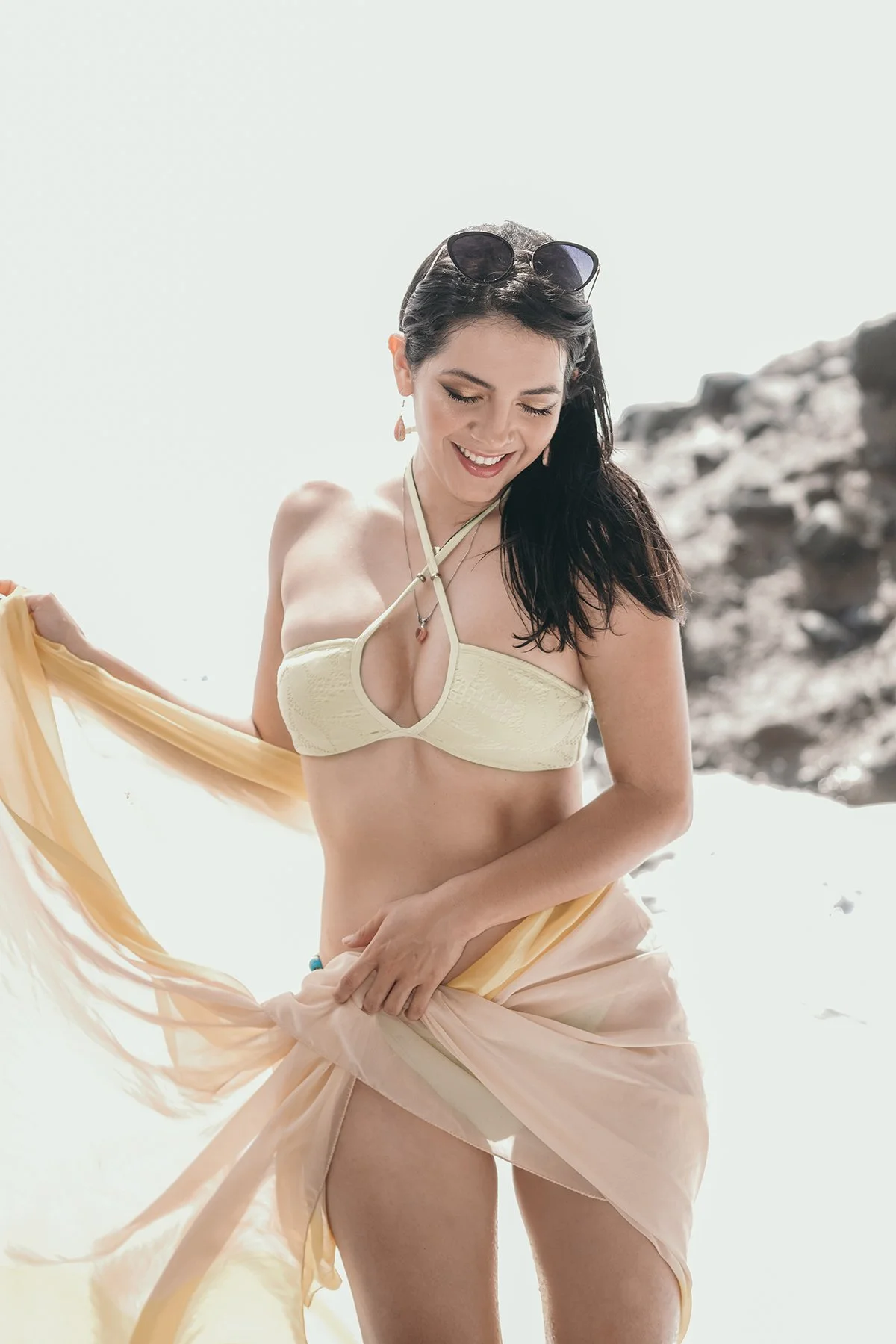 A woman in a yellow bikini top and sheer peach-colored skirt standing in bright sunlight on a beach with rocks in the background, smiling and holding her flowing skirt.