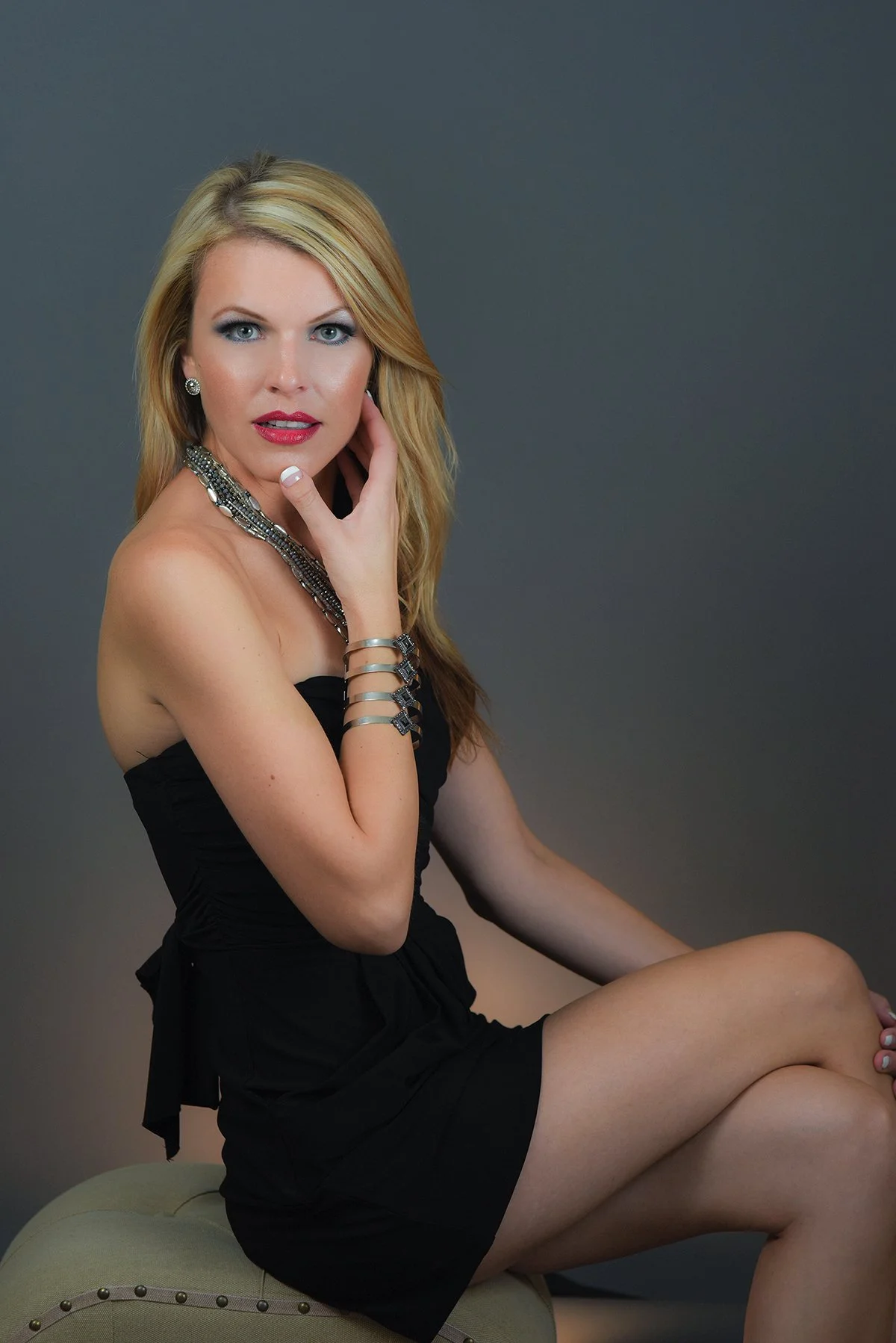 A blonde woman with blue eyes and red lipstick posing in a black strapless dress, sitting on a beige upholstered stool with her hand touching her face, wearing jewelry including earrings, a necklace, and bracelets, against a plain gray background.