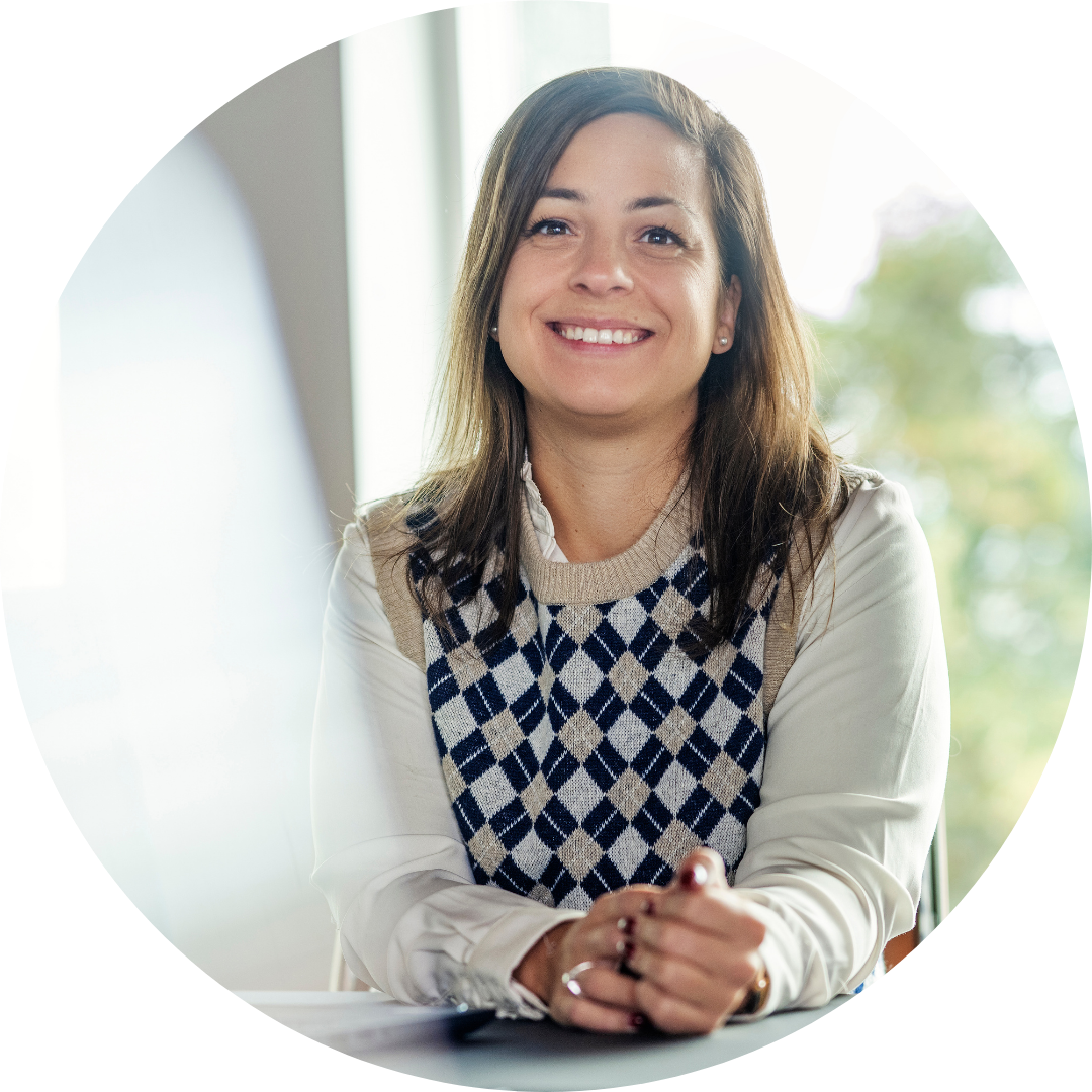 Portrait of a smiling woman with light hair in a white collared shirt and white lab coat with black floral pattern, against a neutral background.