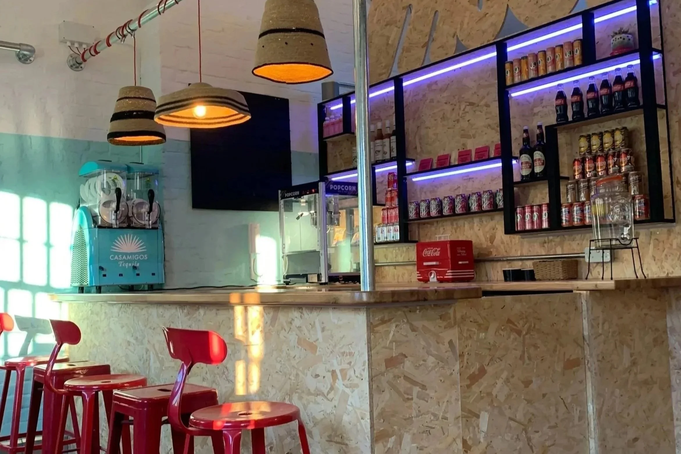 Interior of a modern cafe or bar with high red chairs at a counter, hanging cone-shaped pendant lights, a textured wood wall with illuminated shelves holding soda cans, bottles, and snacks, a large TV screen, and a blue tequila machine.