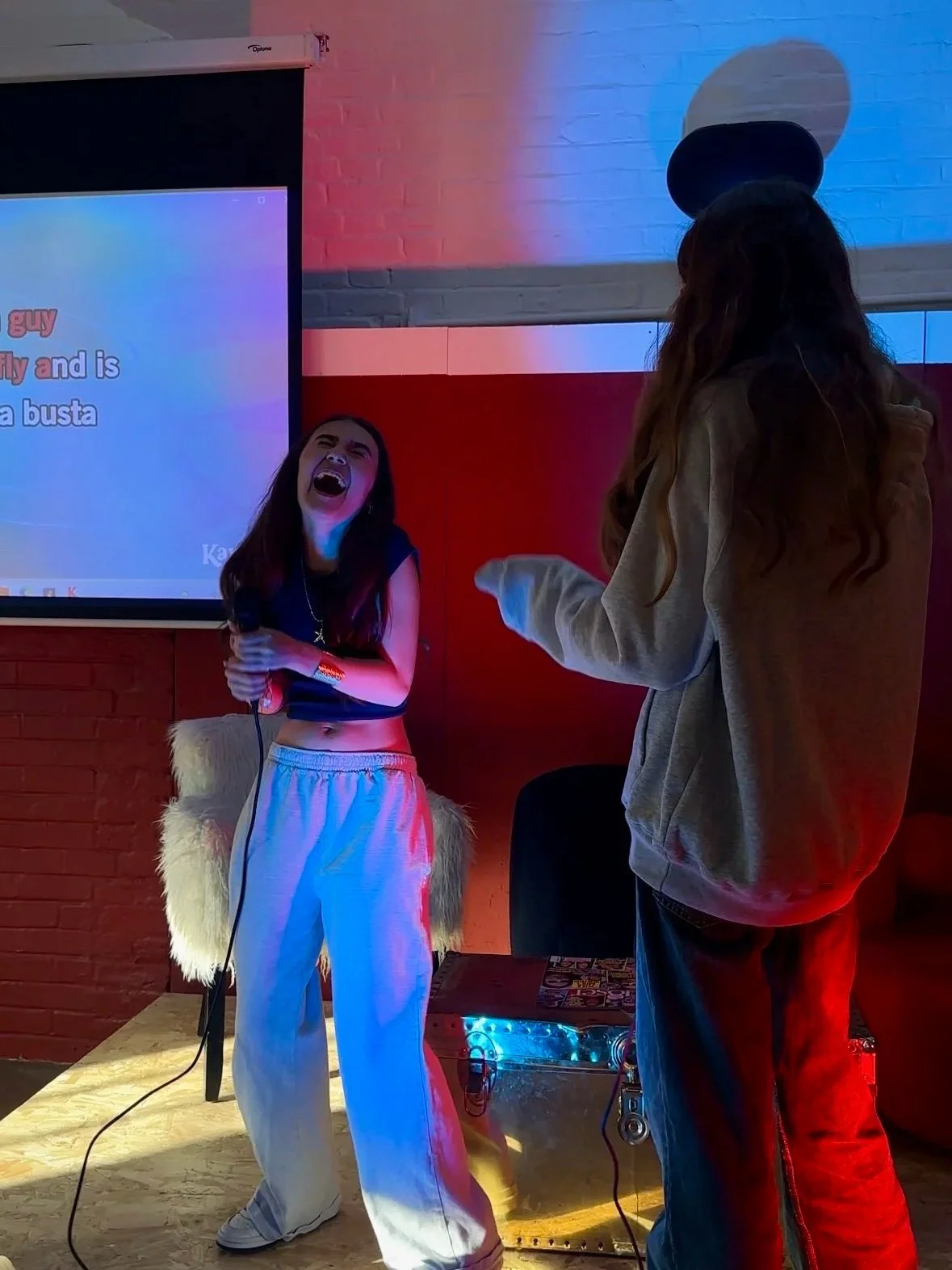 Two women are singing together on a stage with a screen displaying lyrics behind them. One woman, on the left, is holding a microphone and singing with her mouth wide open. She is wearing a sleeveless top and light-colored sweatpants. The other woman, on the right, is facing away from the camera, wearing a hat and a hoodie. The stage has colorful lighting, and a furry chair and an equipment case are visible in the background.