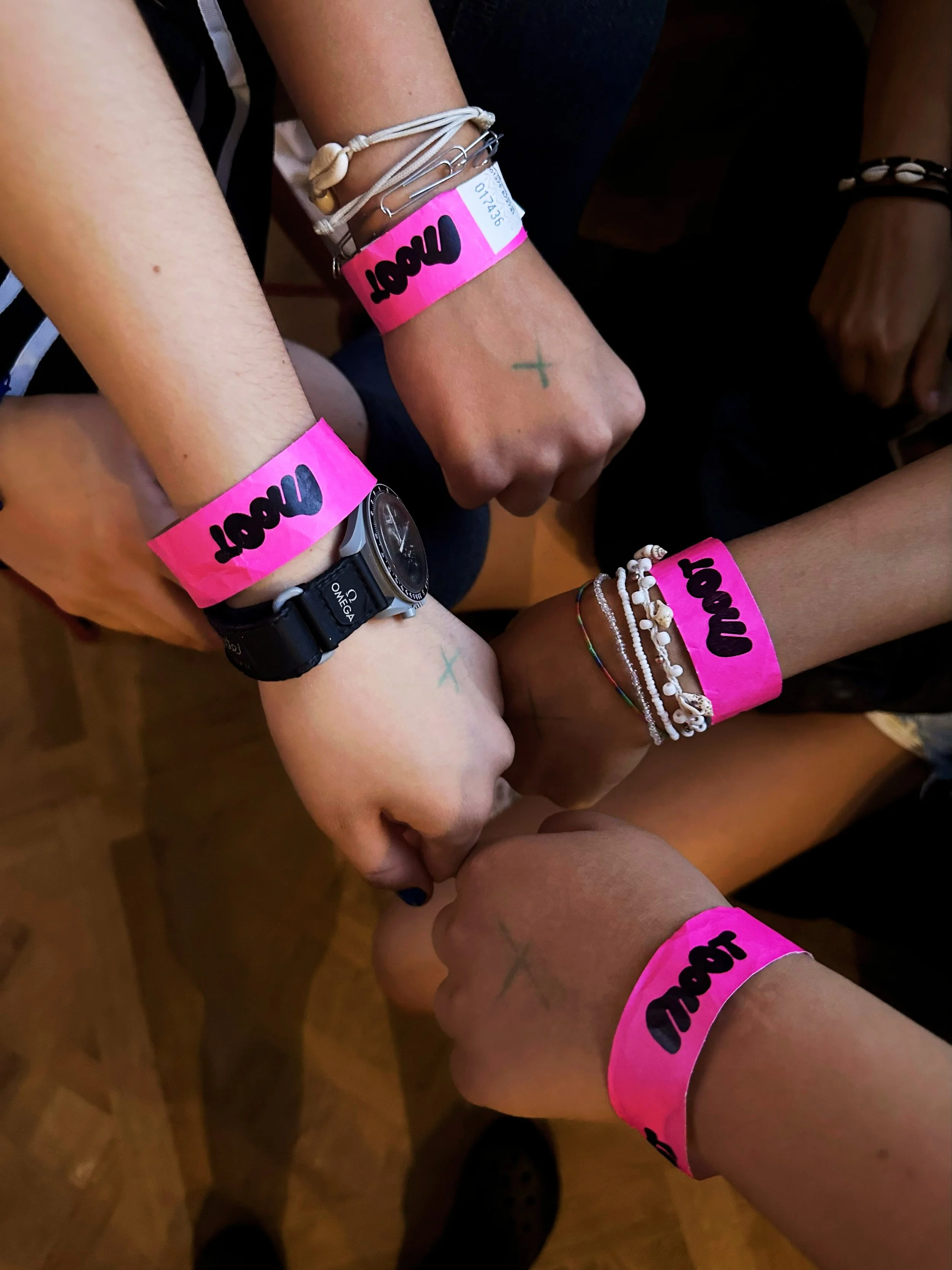 Four people with their fists together, each wearing pink wristbands with the word 'LOVE' written on them and small cross tattoos on their hands. They are also wearing various bracelets and watches.