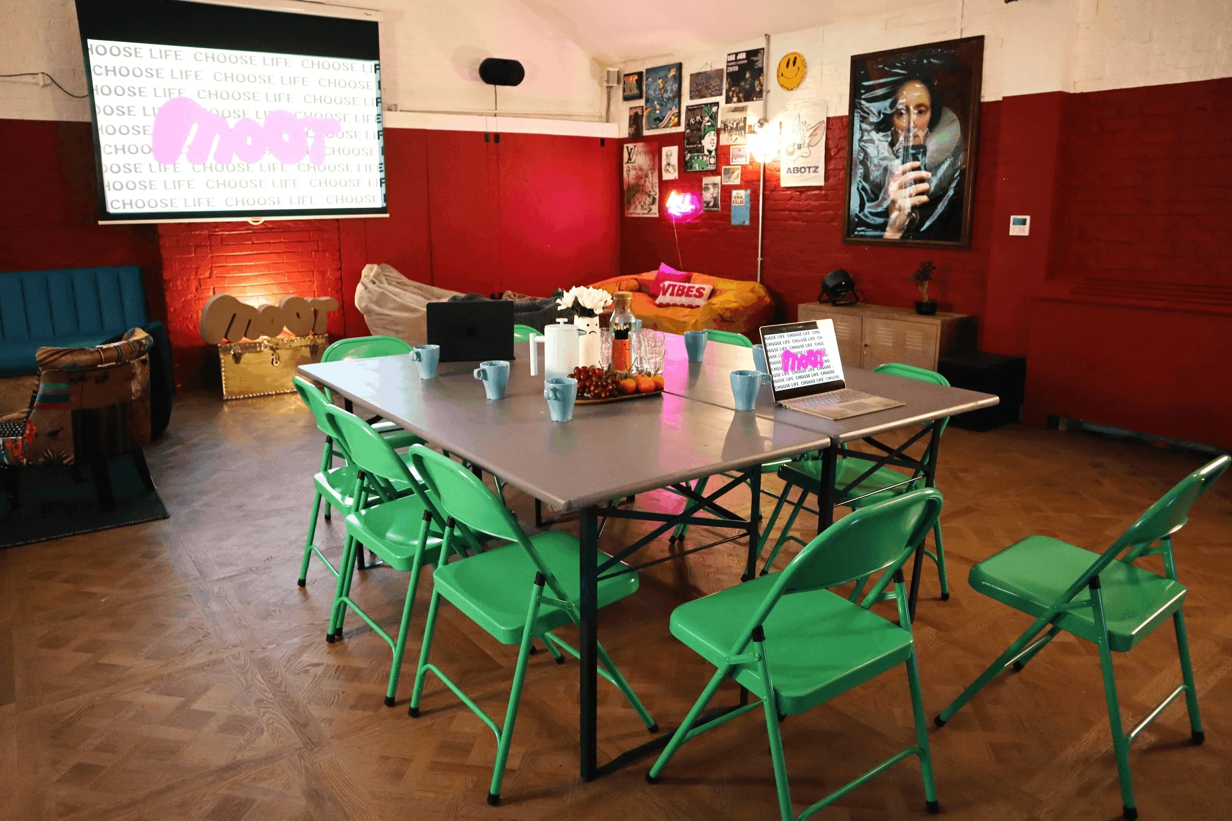 A colorful indoor space with a large table surrounded by green chairs. The table has cups, a laptop, and decorative items. The room features red brick walls, art posters, a large screen displaying 'Choose Life', and wall decorations including a smiley face and framed artwork. There's a cozy seating area with cushions and a neon sign reading 'Love' in the background.