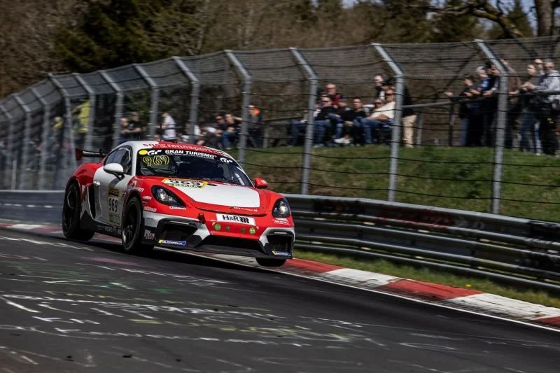 A red and white race car is racing on a track, kicking up dust, with spectators watching behind a fence.