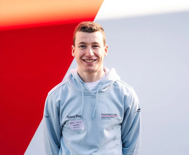 A race car driver sits in their car, wearing a racing suit and helmet, with safety harnesses secured, looking out through the side window with a focused expression.