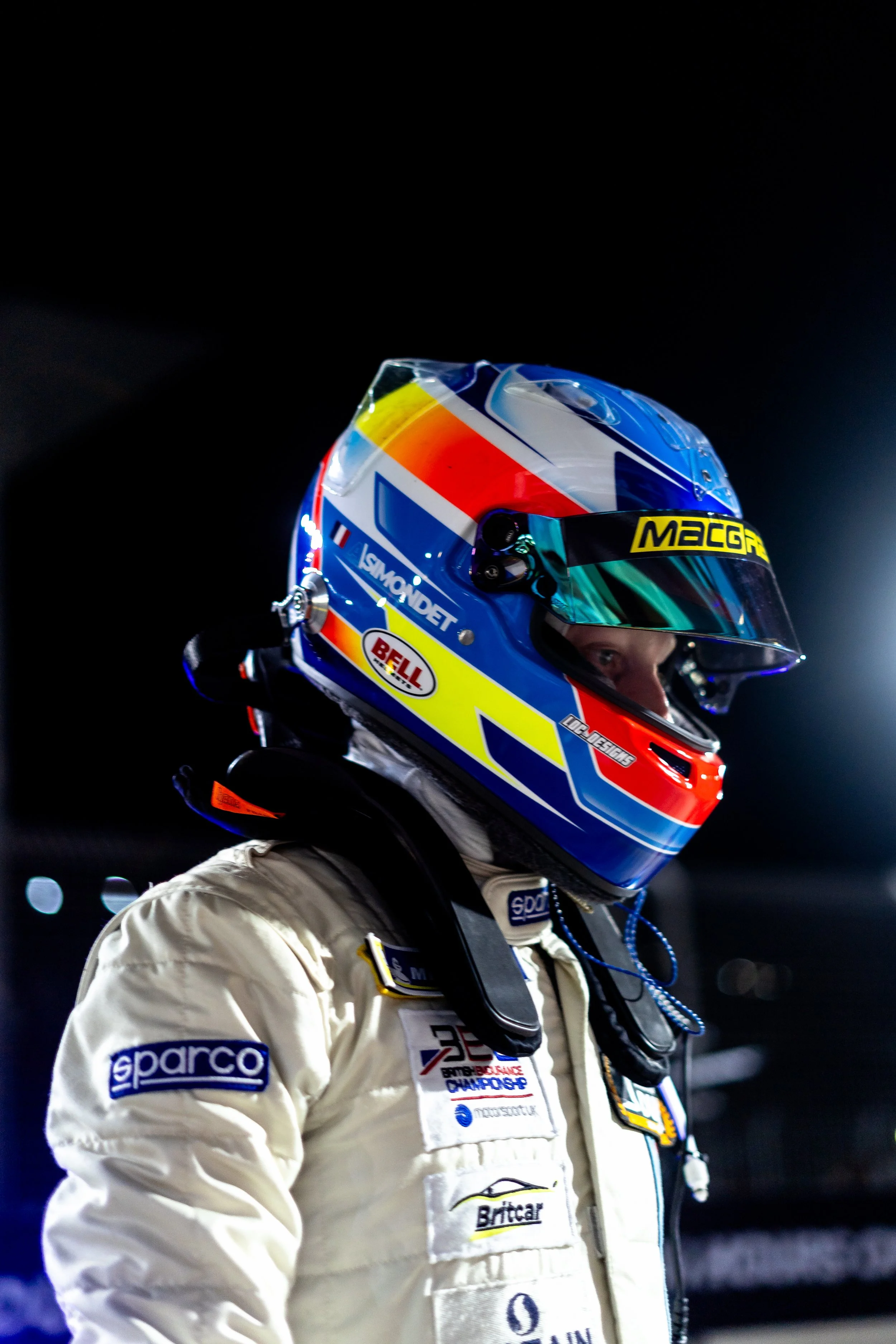 A race car driver wearing a colorful helmet and white racing suit, facing to the right in a dimly lit environment.