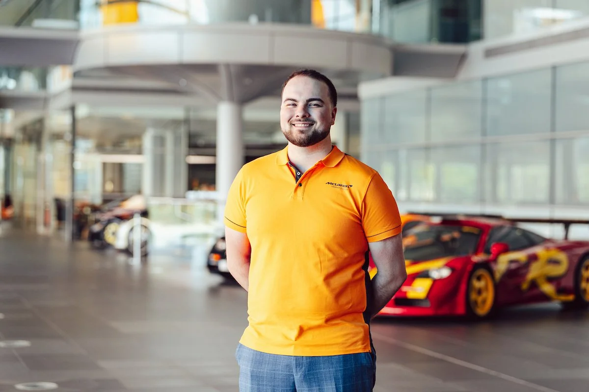 A race car driver wearing a helmet and racing suit, making a peace sign with his hand at a racing event, with a building and other people in the background.