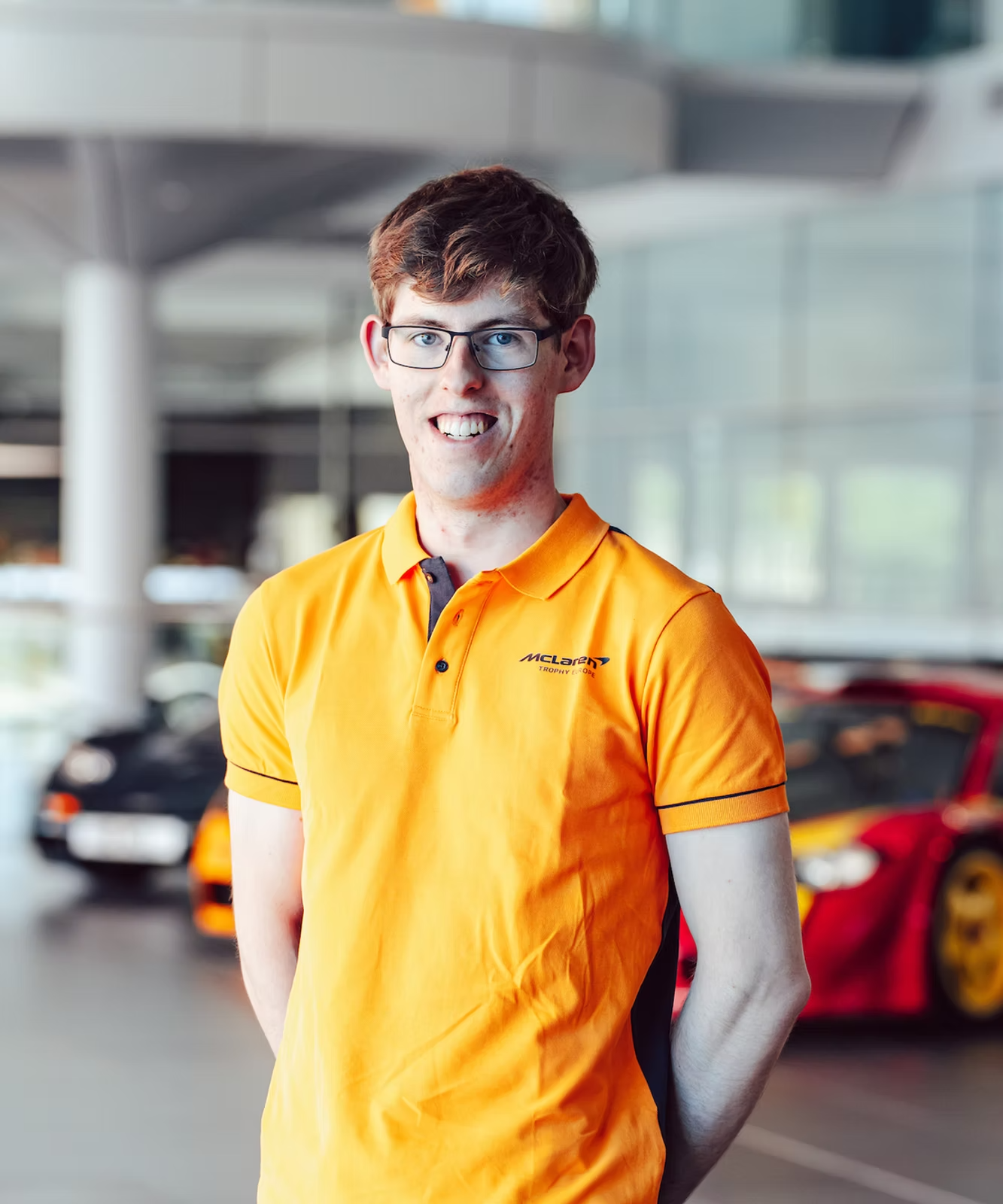 A young man with glasses and short brown hair wearing an orange McLaren Prince of Pace polo shirt, standing indoors with blurred race cars in the background.
