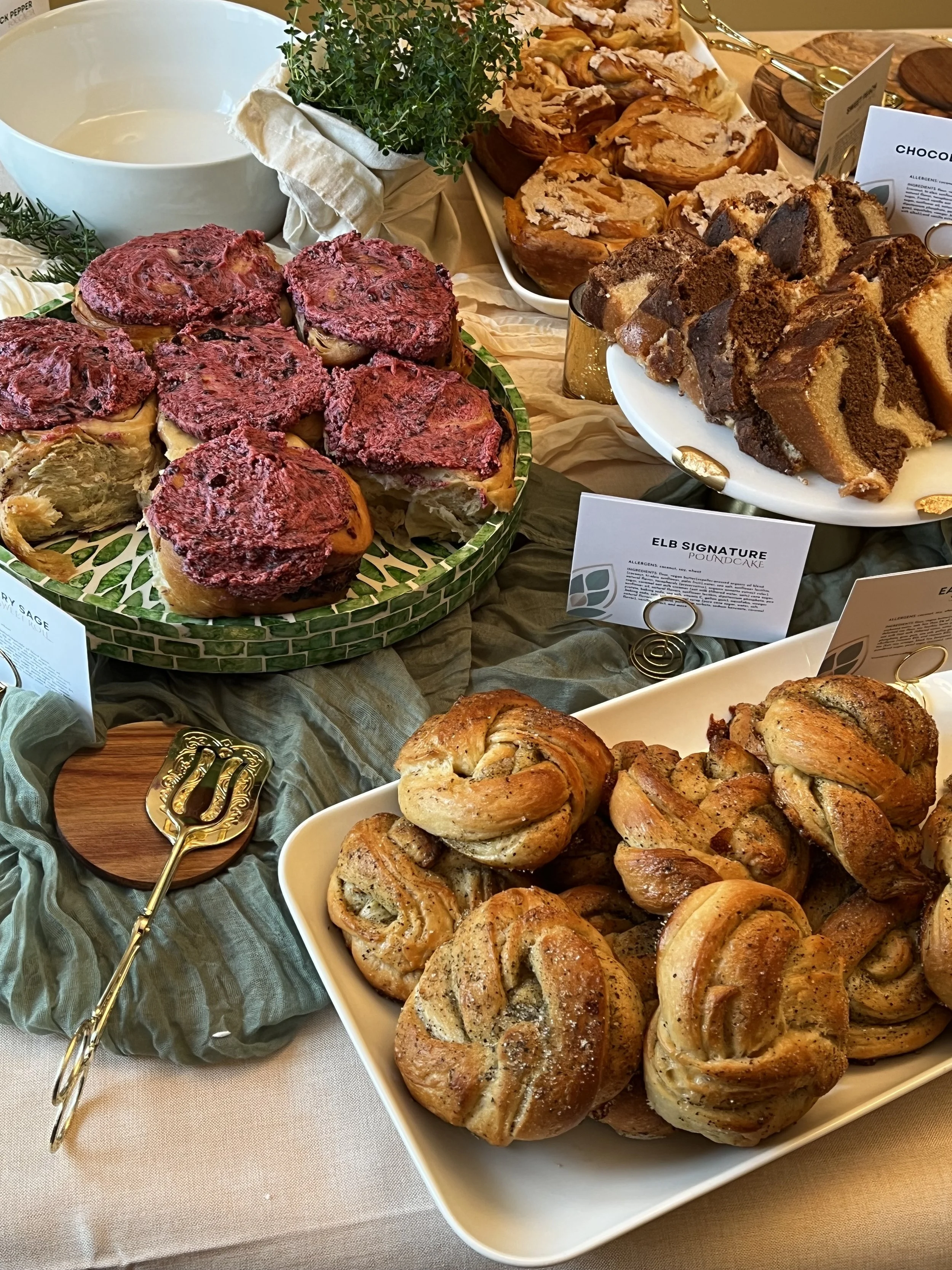 Assorted baked pastries and cakes on display, including cinnamon knots, chocolate swirl cake, and a layered cake with a label, arranged on plates and a table with a green cloth and decorative herbs.