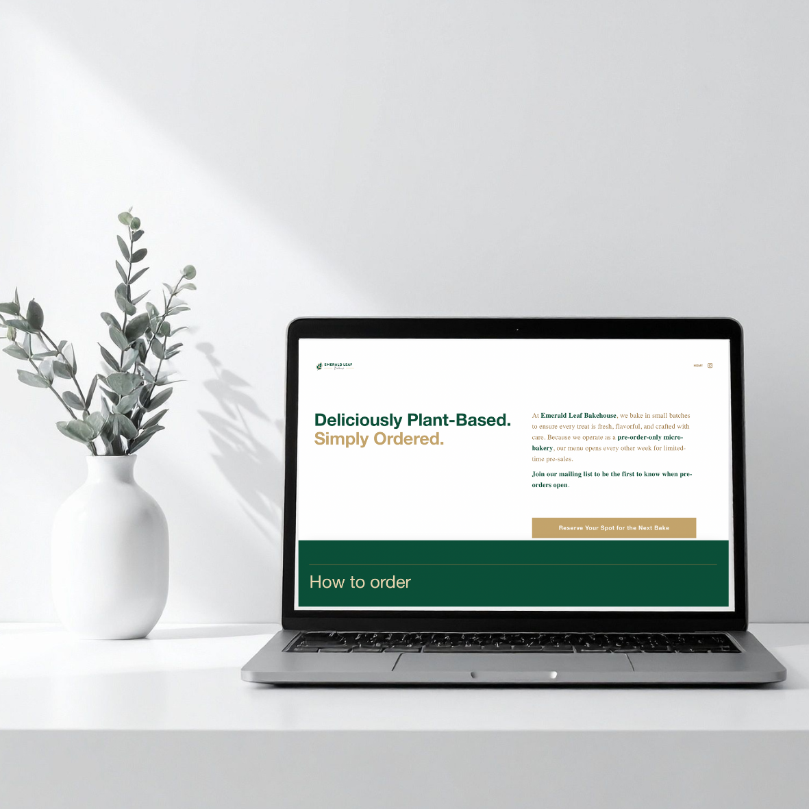 Laptop on white desk displaying a webpage for plant-based baked goods with a minimal white vase with green foliage on the side.