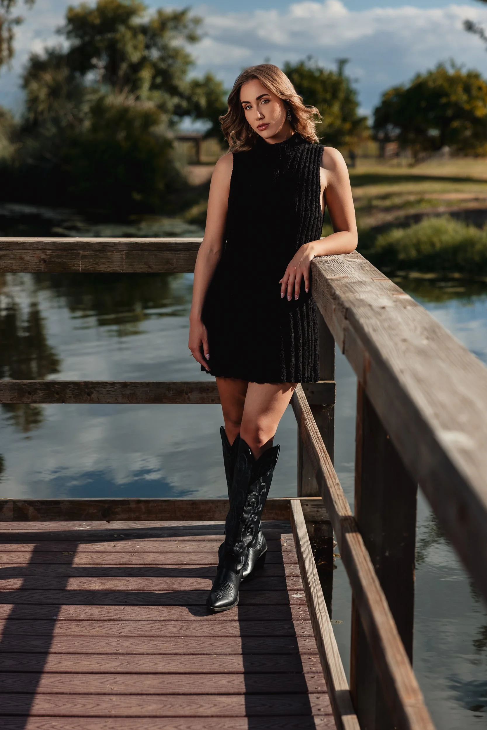 A woman in a black sleeveless dress and cowboy boots posing on a wooden dock beside a body of water with trees and a partly cloudy sky in the background.
