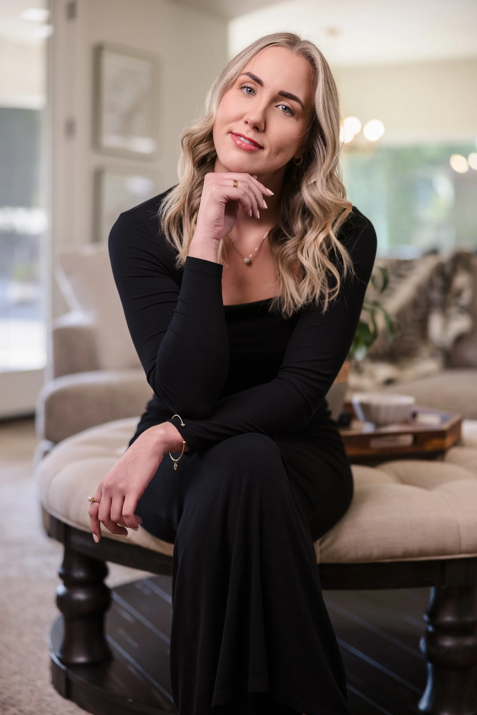 A woman with wavy blonde hair wearing a black long-sleeve top and black pants sitting on a beige cushioned bench in a well-lit room, smiling and resting her chin on her hand.