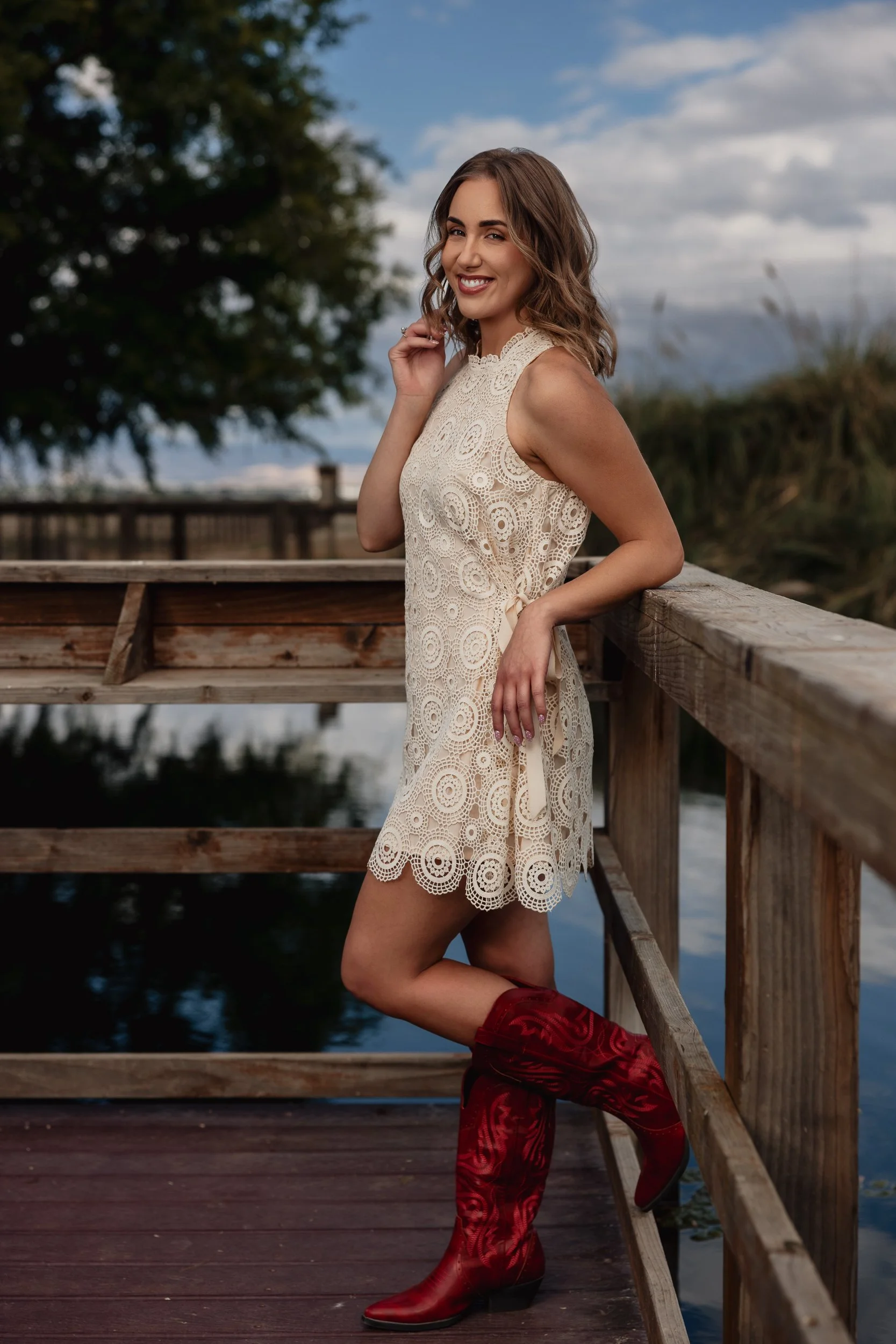 A woman in a cream lace dress and red cowboy boots leaning against a wooden railing on a dock near water, with a tree and cloudy sky in the background.