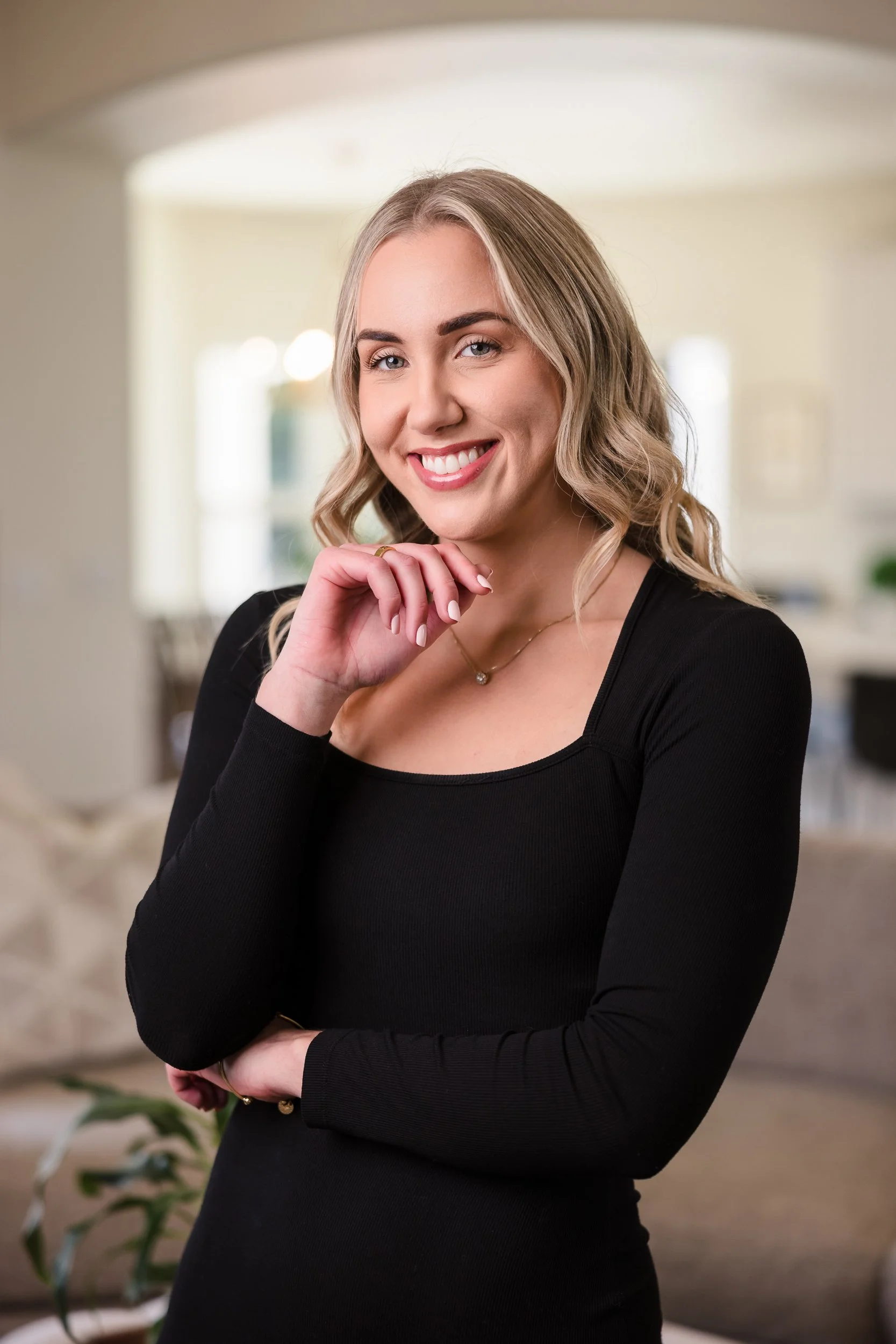 A smiling woman with blonde, wavy hair wearing a black long-sleeve top, standing indoors with a bright, blurred background.