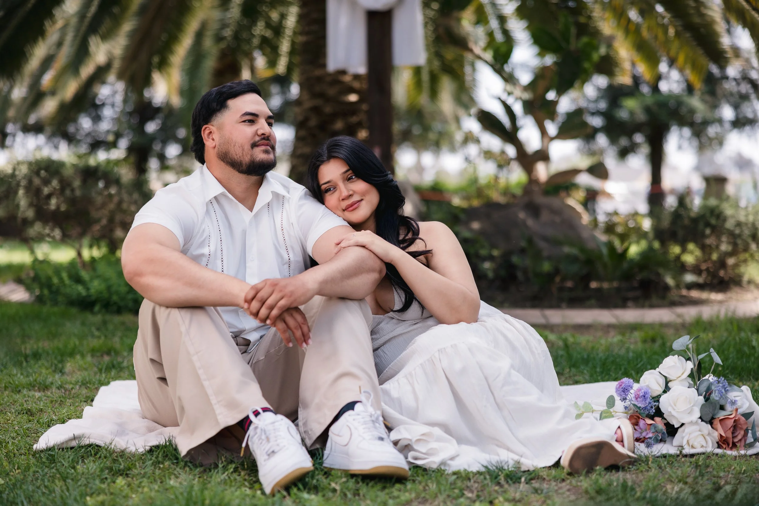 A couple sitting on a blanket in a park, surrounded by trees, with flowers nearby. The woman is resting her head on the man's shoulder, and they are both looking content and relaxed.