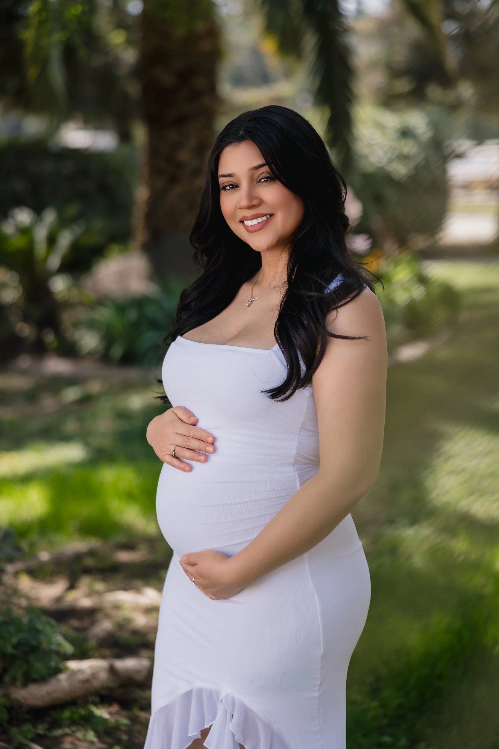 A pregnant woman with long black hair smiling outdoors, wearing a white dress in a park with trees and greenery in the background.