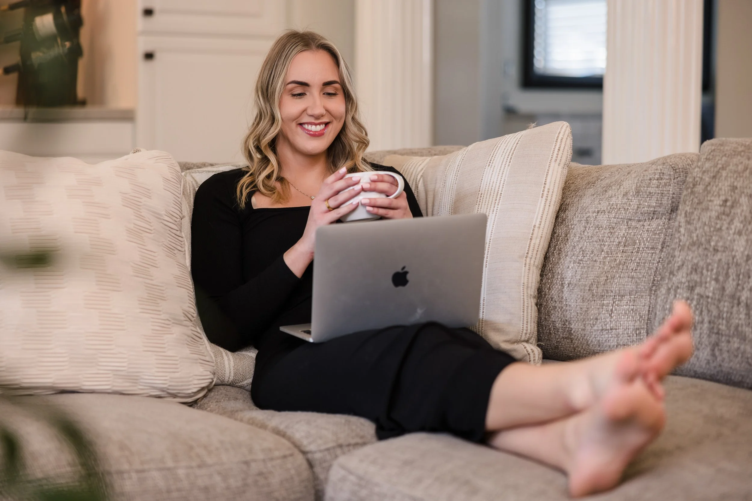 A woman sitting on a beige couch, smiling, holding a cup, using a laptop, in a well-lit living room.