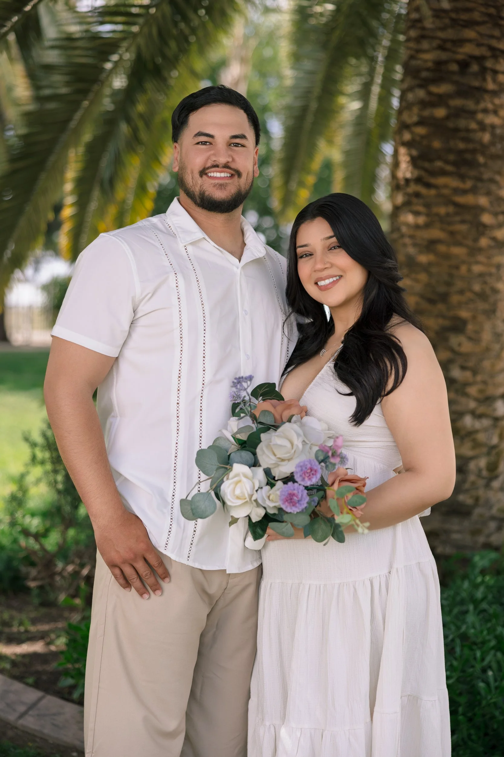 A couple standing outdoors under palm trees, dressed in light-colored clothing, smiling. The woman is holding a bouquet of white, purple, and pink flowers with eucalyptus leaves.