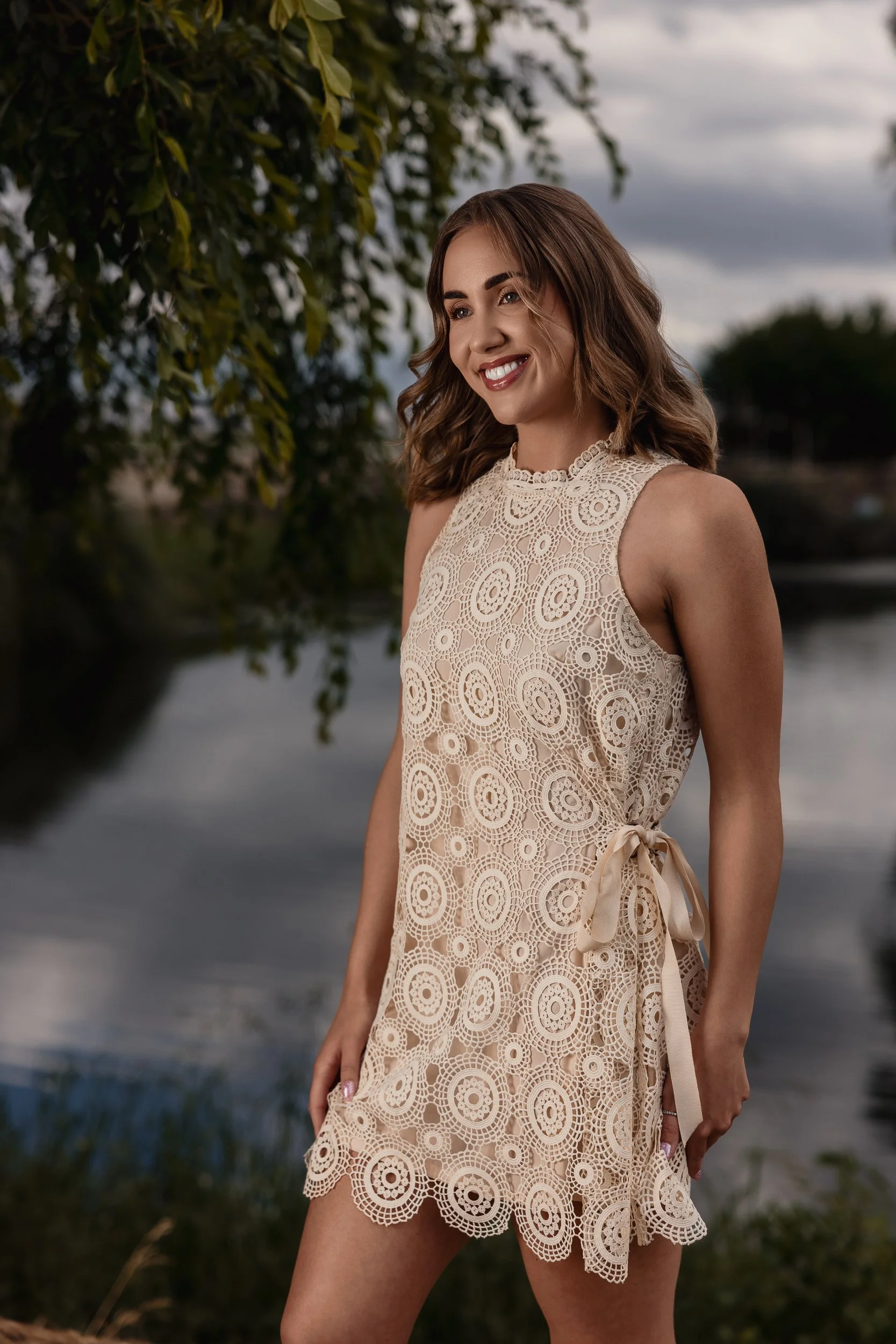 A woman in a sleeveless, beige lace dress with a ribbon tied at the waist stands outdoors near water, smiling with trees and cloudy sky in the background.