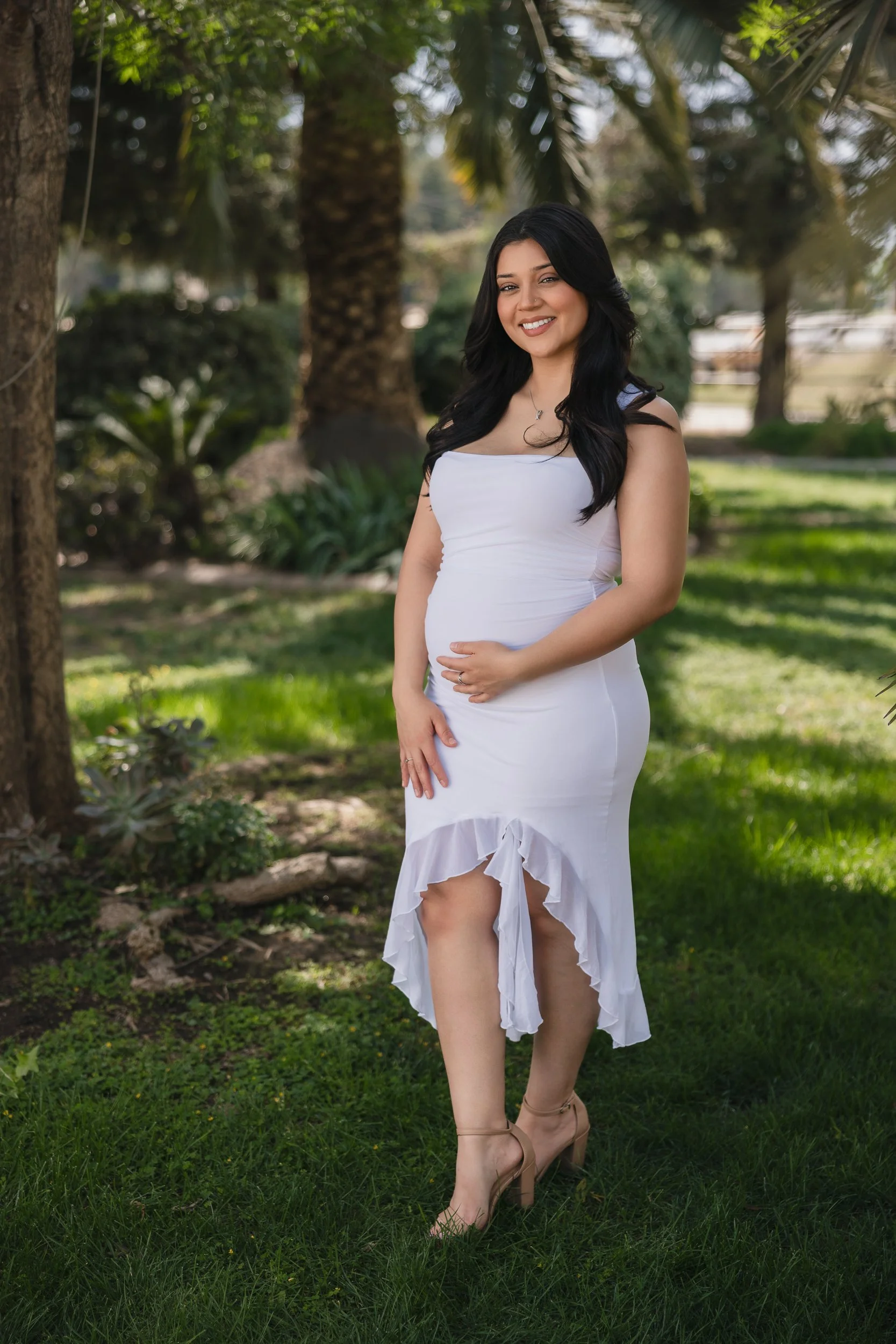 A woman with long dark hair wearing a white dress with ruffles and high heels, standing outdoors on a grassy area with trees in the background, smiling at the camera.
