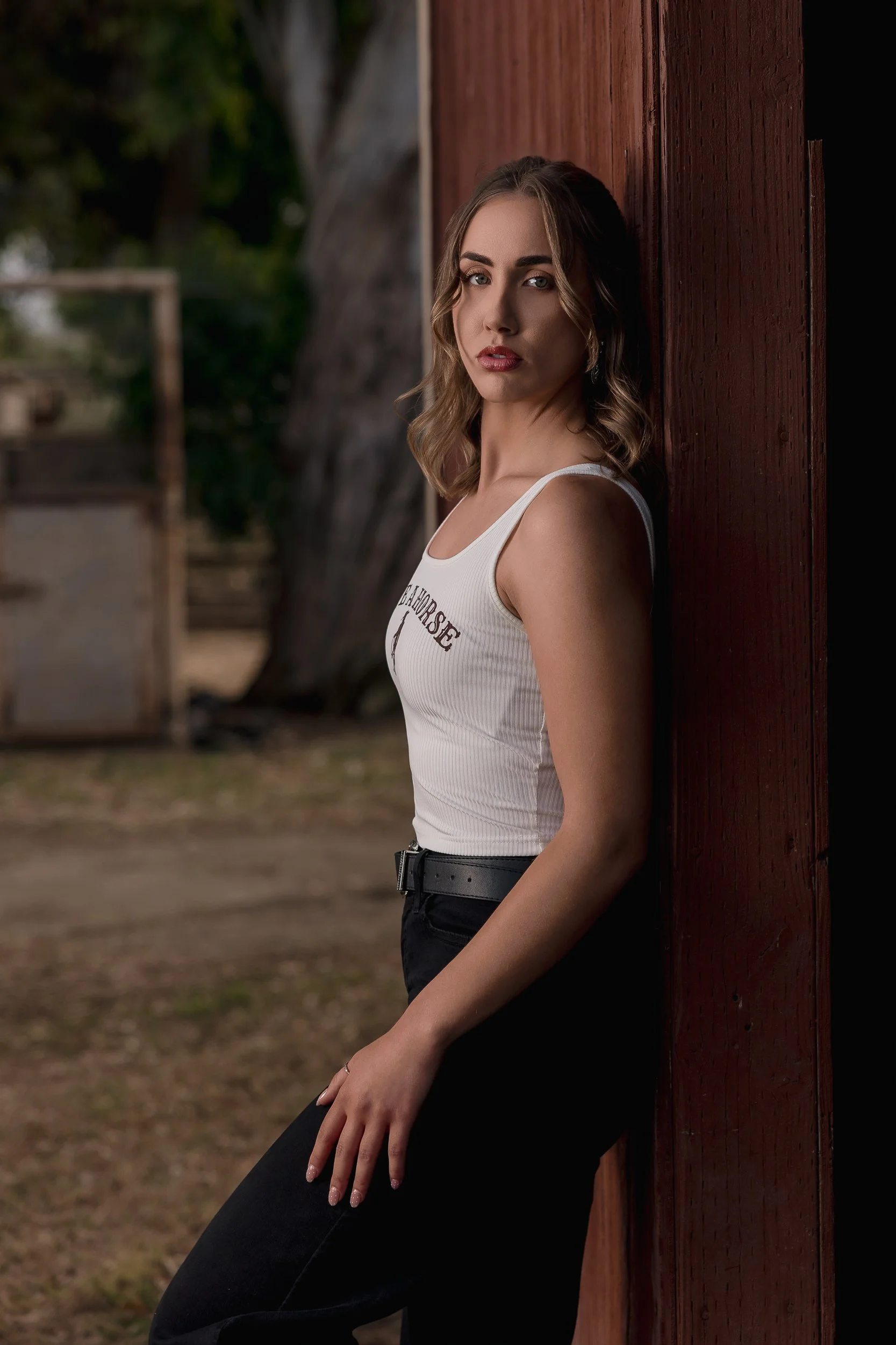 A woman leaning against a wooden fence outdoors, with trees and blurred background.