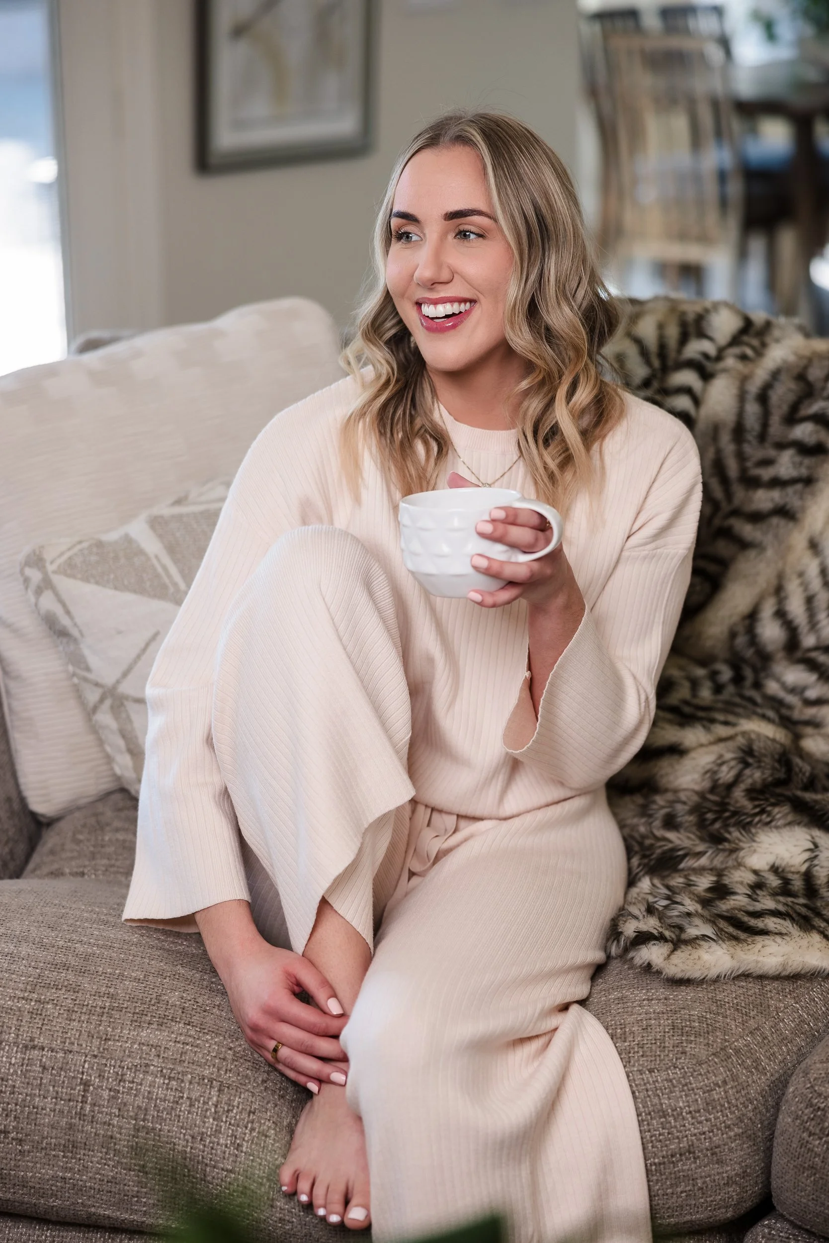A young woman with blonde, wavy hair and light skin smiling while sitting on a beige couch in a cozy living room, holding a white mug. She is wearing a cream-colored sweater and matching loungewear. There is a patterned pillow behind her and a furry 