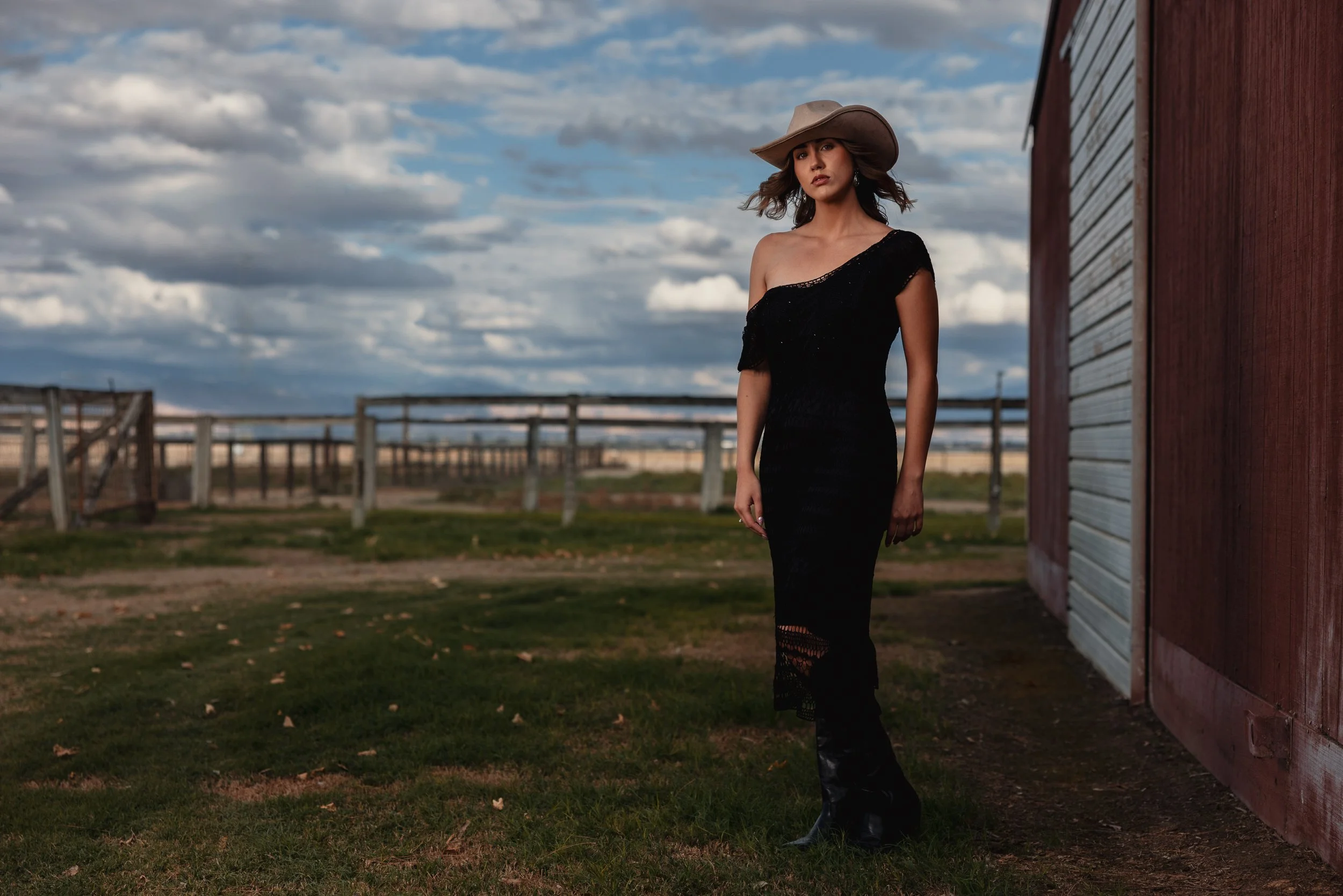 A woman wearing a black dress and a large hat standing outside near a barn on a partly cloudy day.