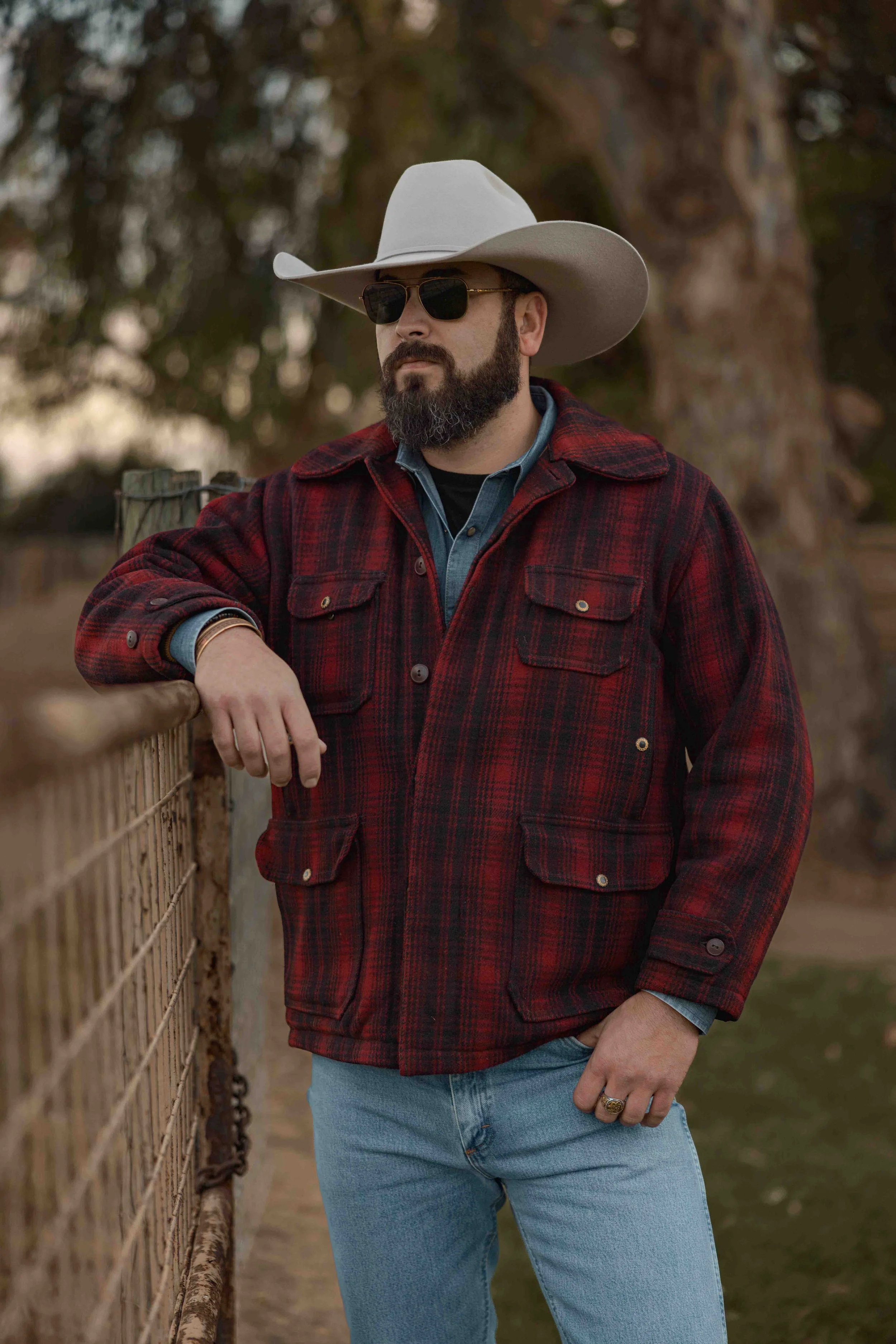 A man with a beard and sunglasses wearing a white cowboy hat, red flannel jacket, and jeans leaning on a wooden fence outdoors.