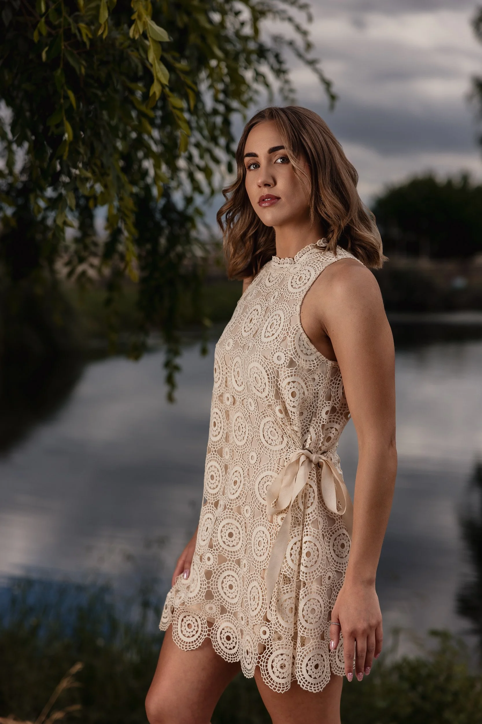 A woman with shoulder-length brown hair and fair skin standing outdoors next to a tree with green leaves, near a body of water under a cloudy sky, wearing a sleeveless beige lace dress with a ribbon tied at the waist.