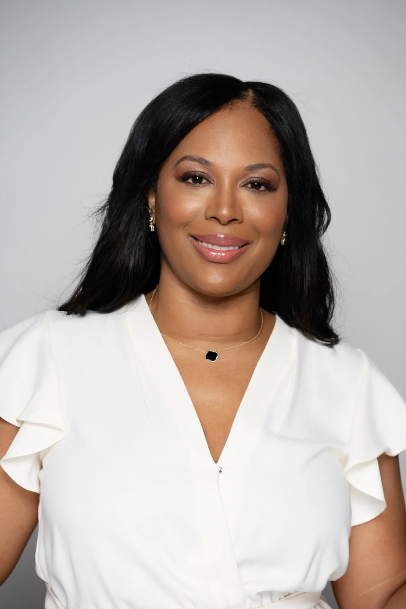 Portrait of a woman with medium skin, black hair, wearing a white top, gold earrings, and a delicate necklace, smiling against a light gray background.