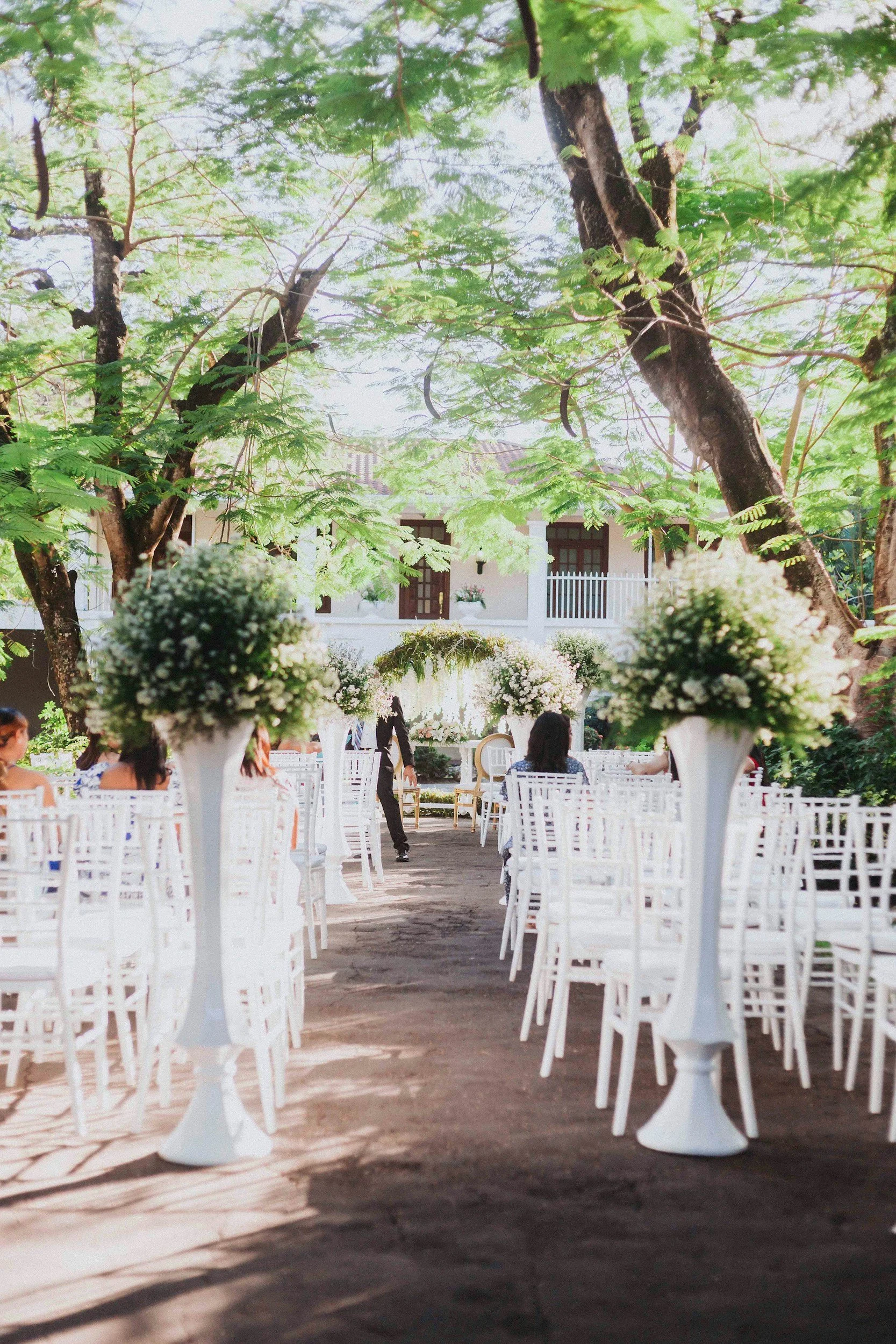 Outdoor wedding ceremony setup with white chairs, floral arrangements, and trees providing shade.