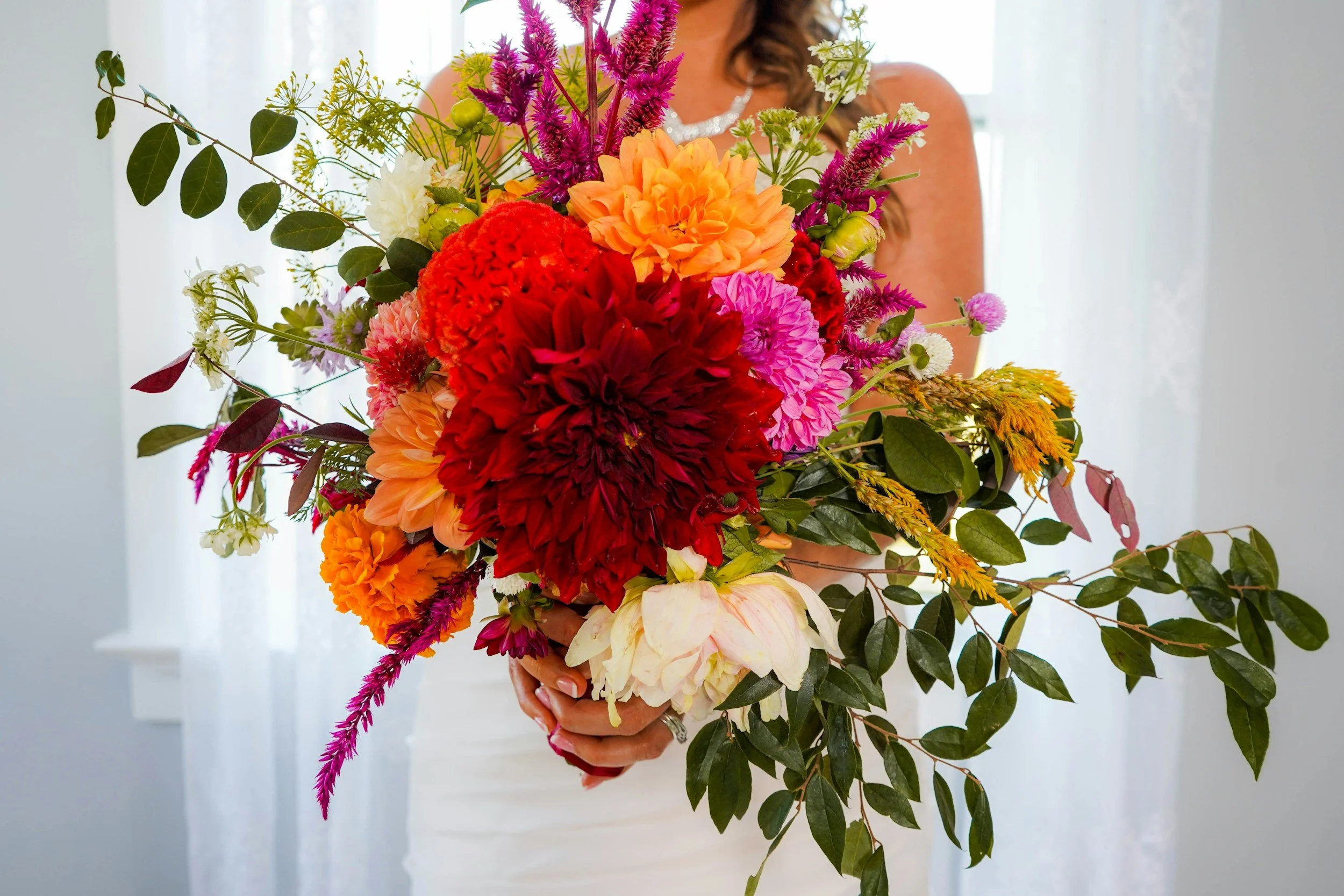 Person holding a large colorful bouquet of various flowers including dahlias, cockscomb, and greenery, with a light background.