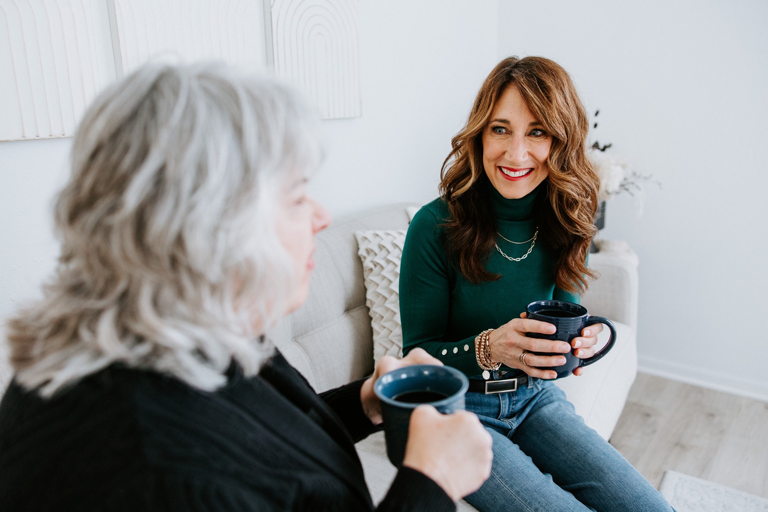 Dana Cushing holding coffee, sitting on white couch, talking to a menopause health client